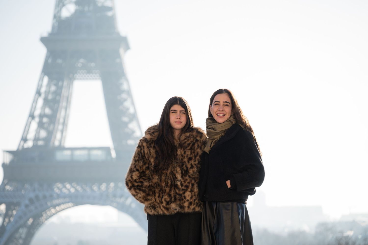 Two people pose together in winter coats with the iconic Eiffel Tower visible in the misty Paris background.