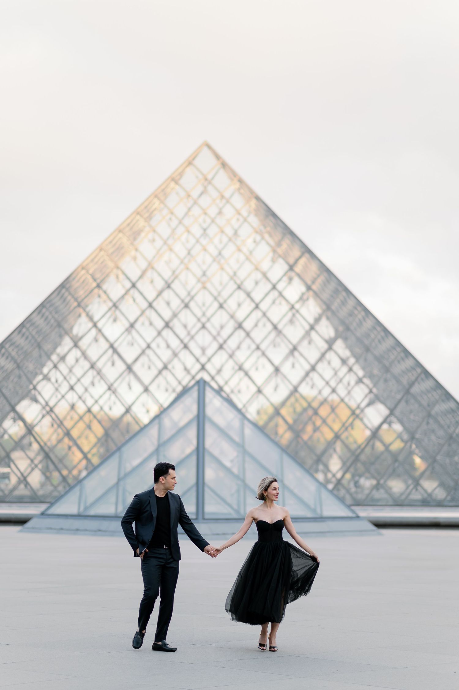 Couple holding hands in front of the Louvre Pyramid in Paris, woman in black tulle dress, man in dark suit.