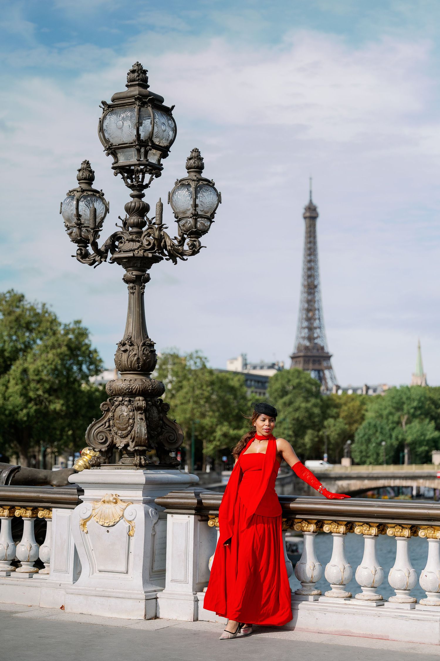 Woman in red gown posing by ornate lamppost on Pont Alexandre III bridge with Eiffel Tower in Paris background.
