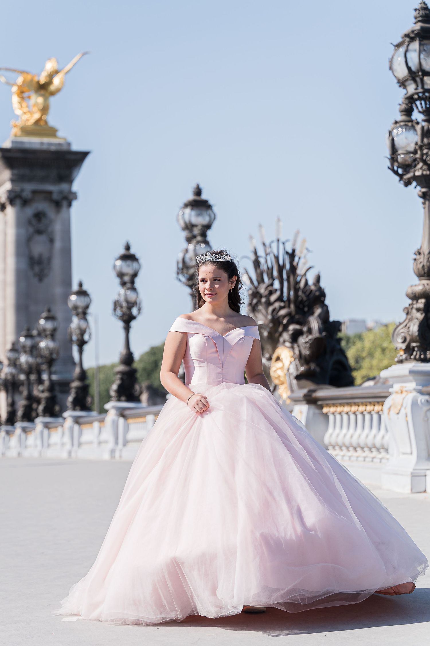 Young woman in stunning pink off-shoulder ball gown with tiara posing on Pont Alexandre III bridge in Paris.