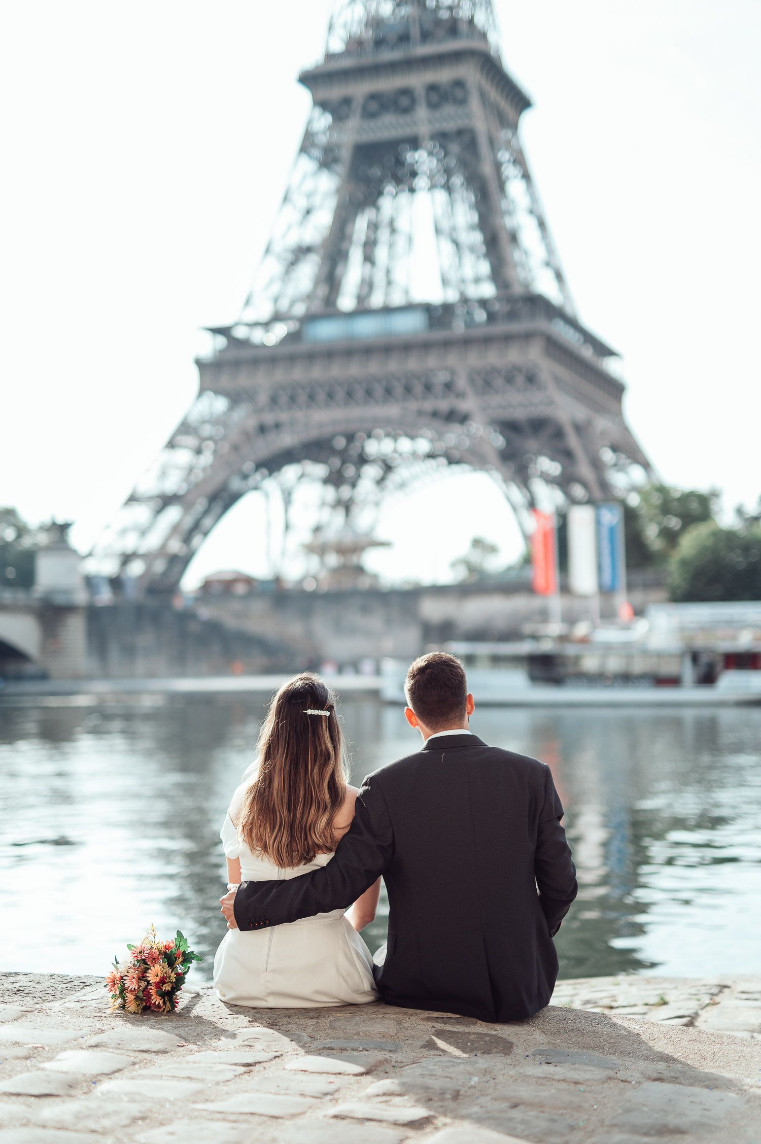 A romantic couple sits by the Seine River overlooking the iconic Eiffel Tower in Paris.