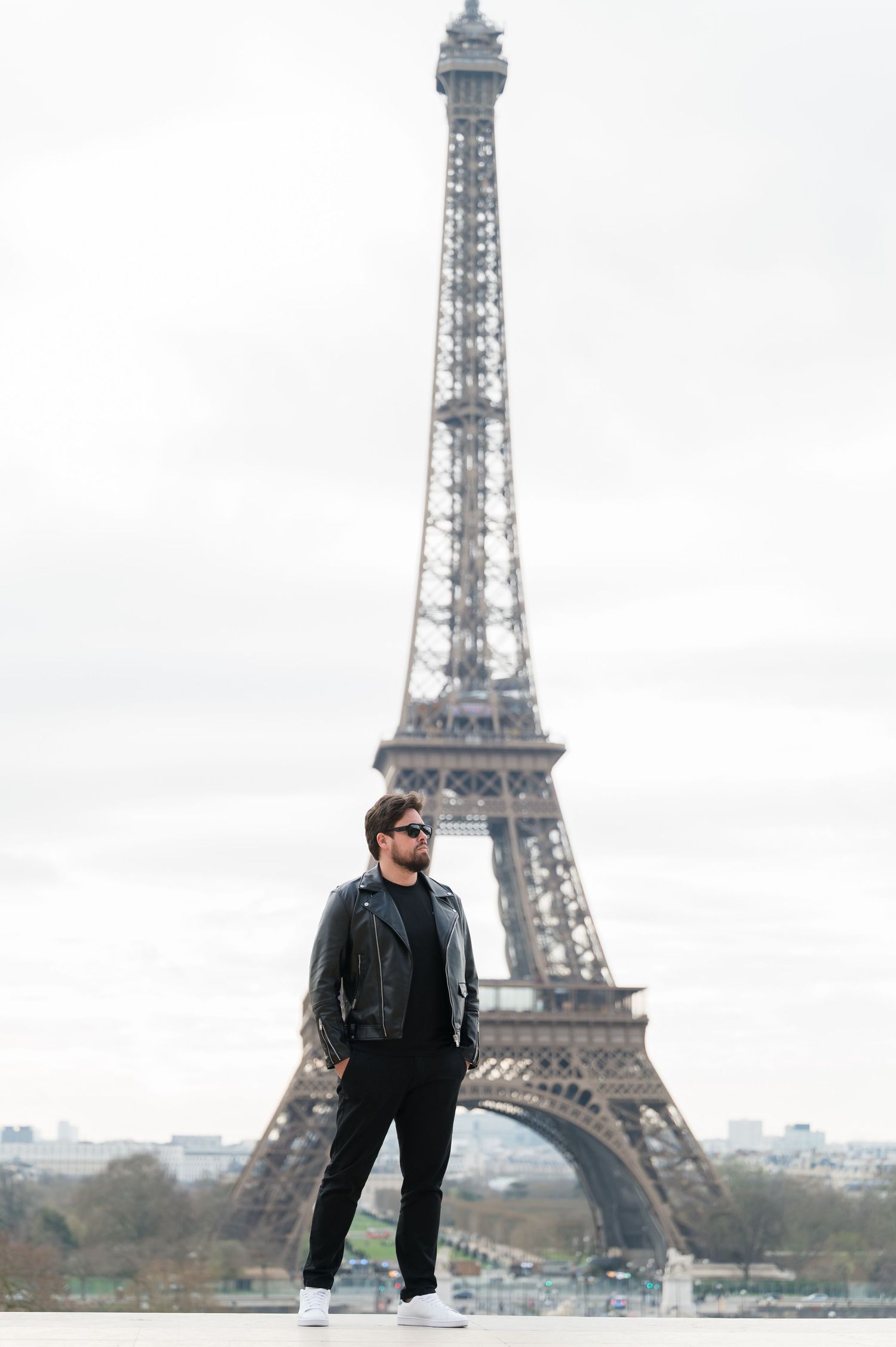 A stylish man in a black leather jacket and sunglasses stands confidently in front of the Eiffel Tower in Paris.