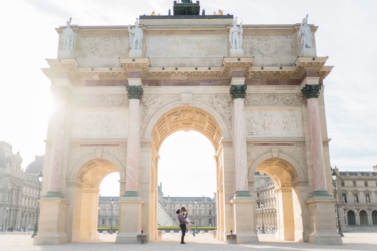 Couple embracing under the Arc de Triomphe du Carrousel in Paris, France, with sunlight streaming through the arch.