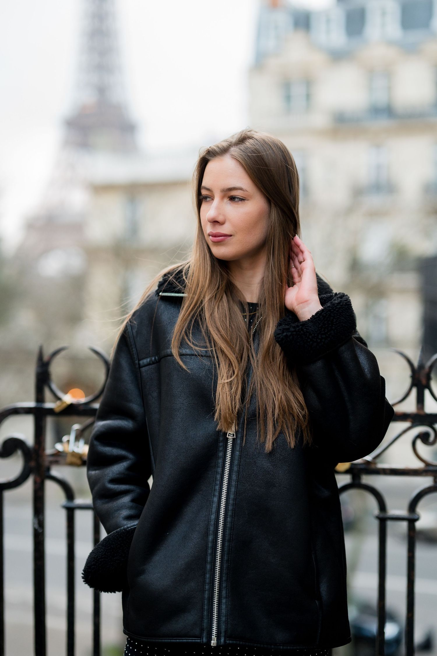 Person in black coat standing by ornate iron railing with Eiffel Tower visible behind.