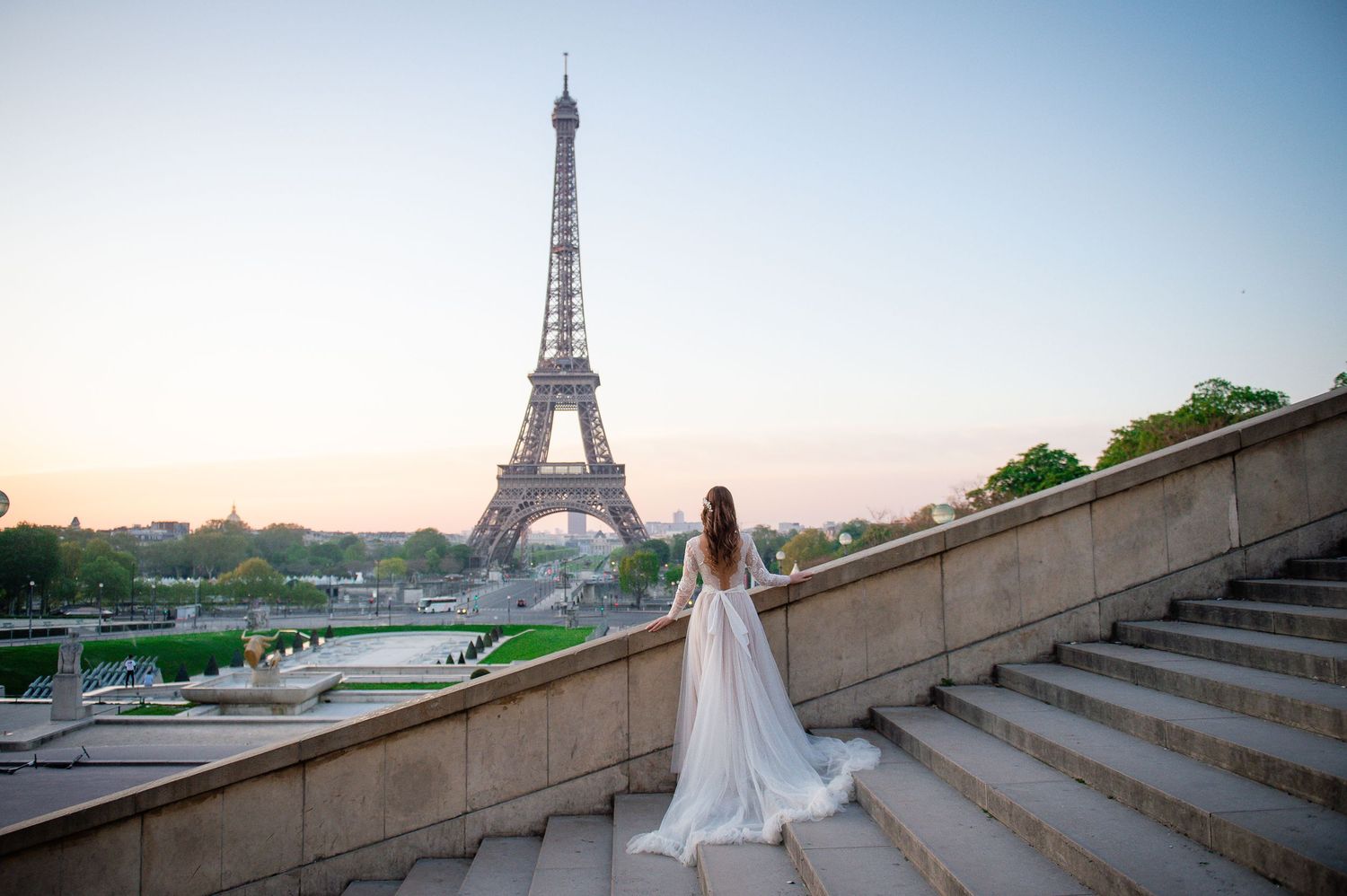 Bride in white gown standing on stone steps with Eiffel Tower backdrop.
