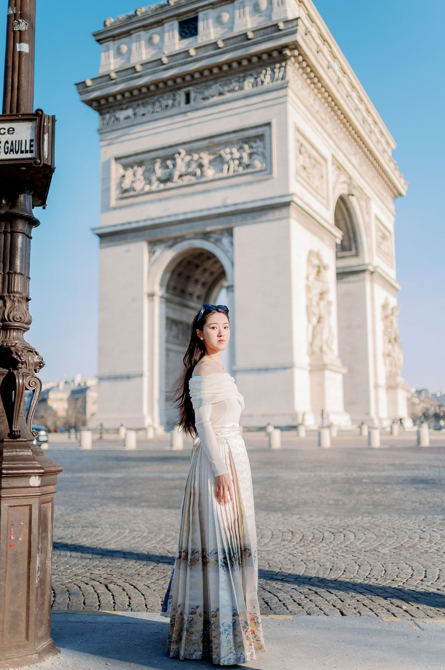 A elegant figure in a flowing dress poses in front of the Arc de Triomphe in Paris.
