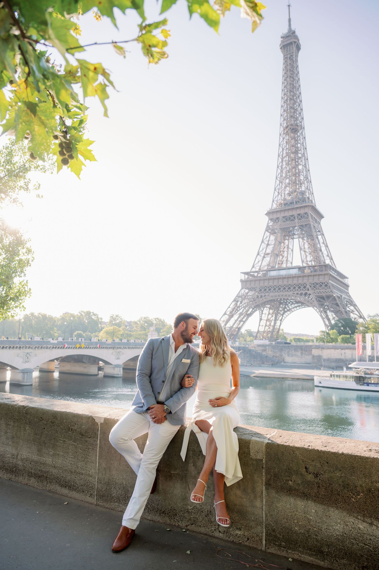 Couple sharing a romantic kiss on a stone wall with the Eiffel Tower and Seine River in Paris background.