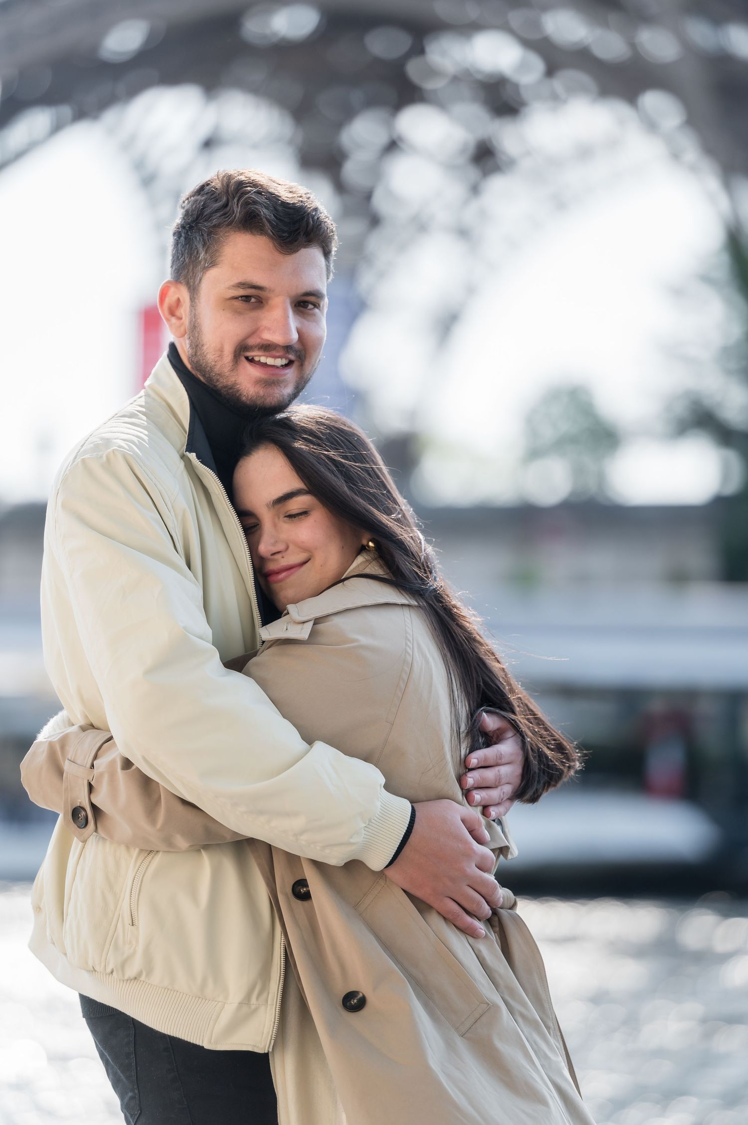 Happy couple sharing a warm hug beneath the Eiffel Tower in Paris, dressed in matching beige trench coats.
