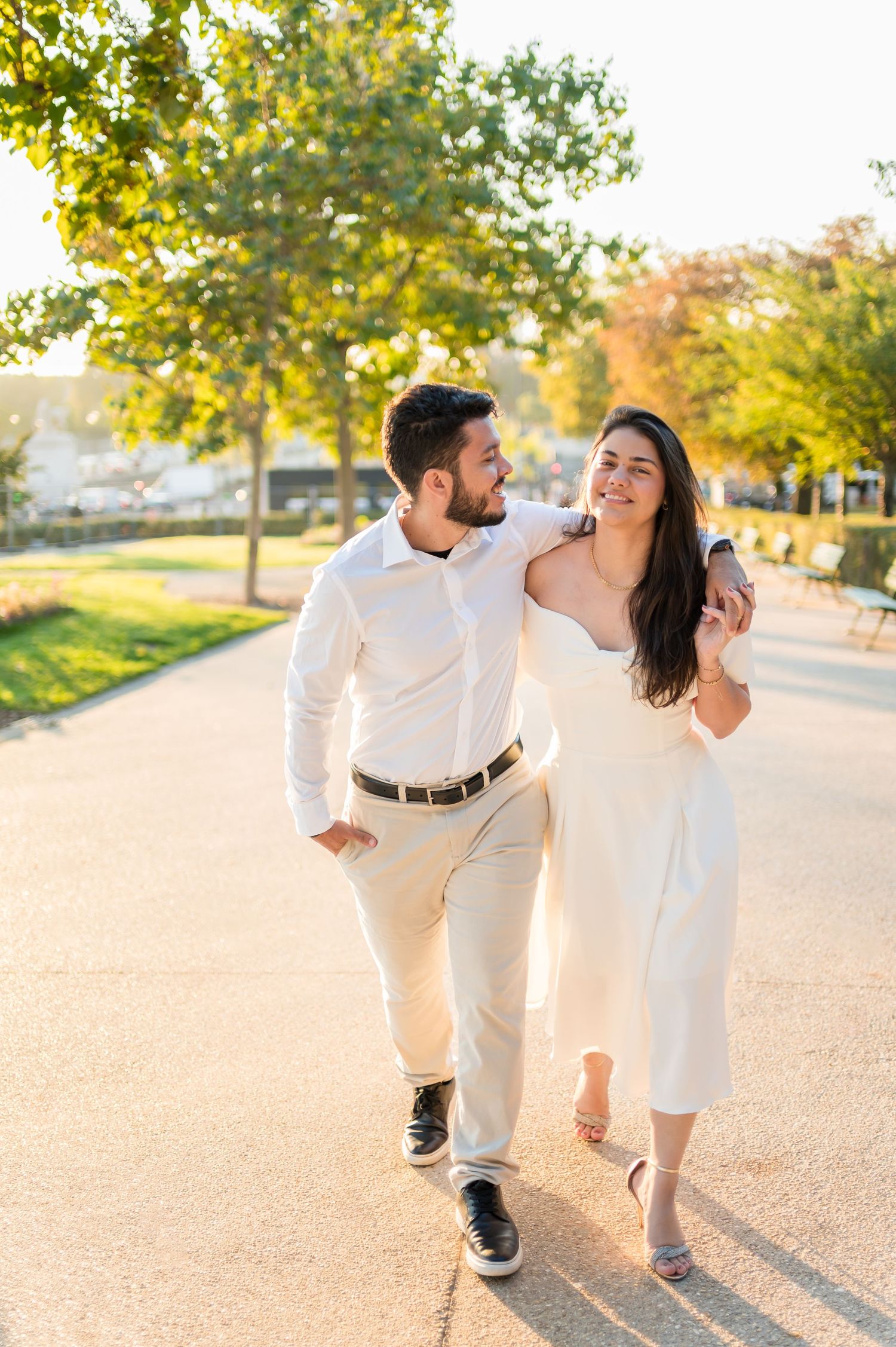 A couple in white outfits dances playfully on a tree-lined path during golden hour.