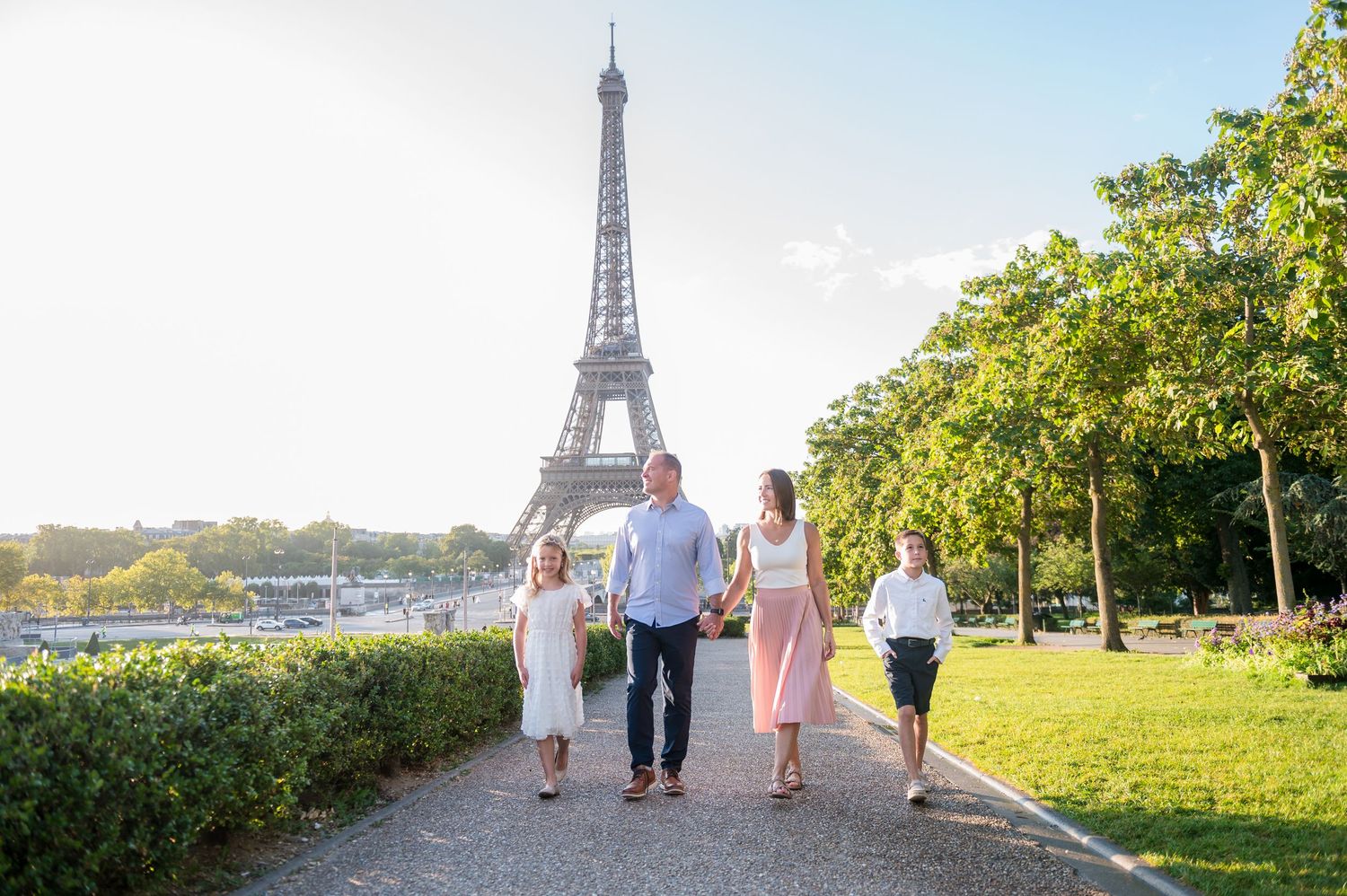 Family walking along tree-lined path with iconic Eiffel Tower in background on sunny summer day in Paris.