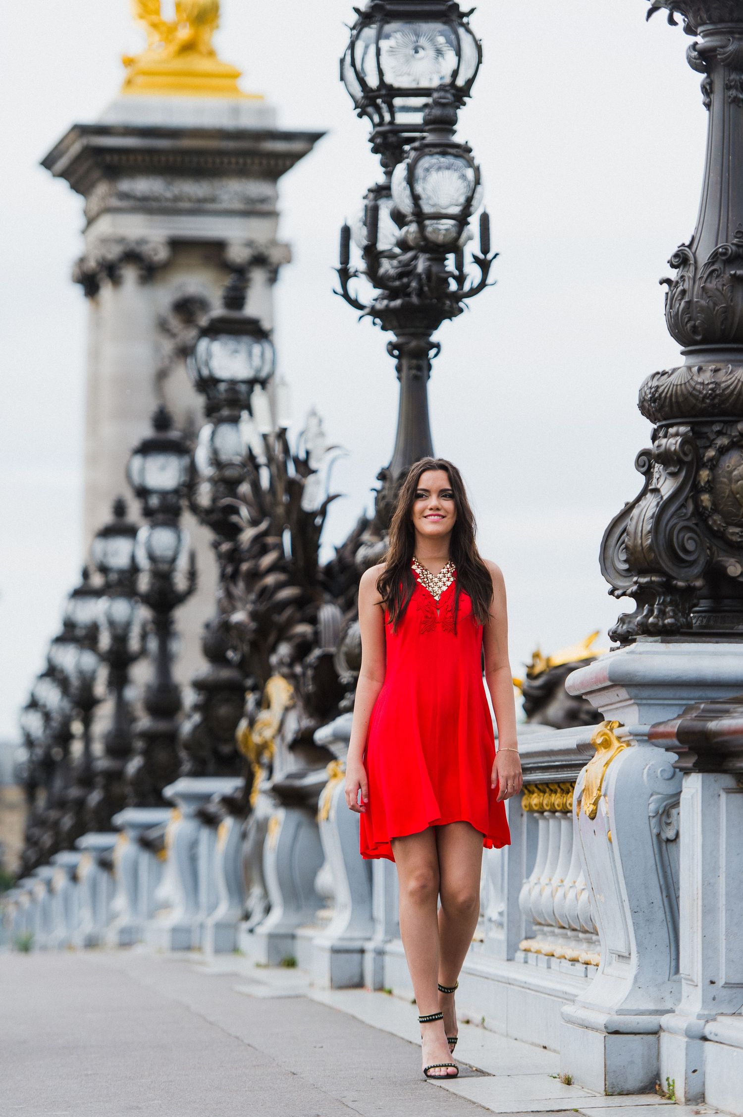 Woman in flowing red dress walking along ornate bridge with classic lampposts in Paris.