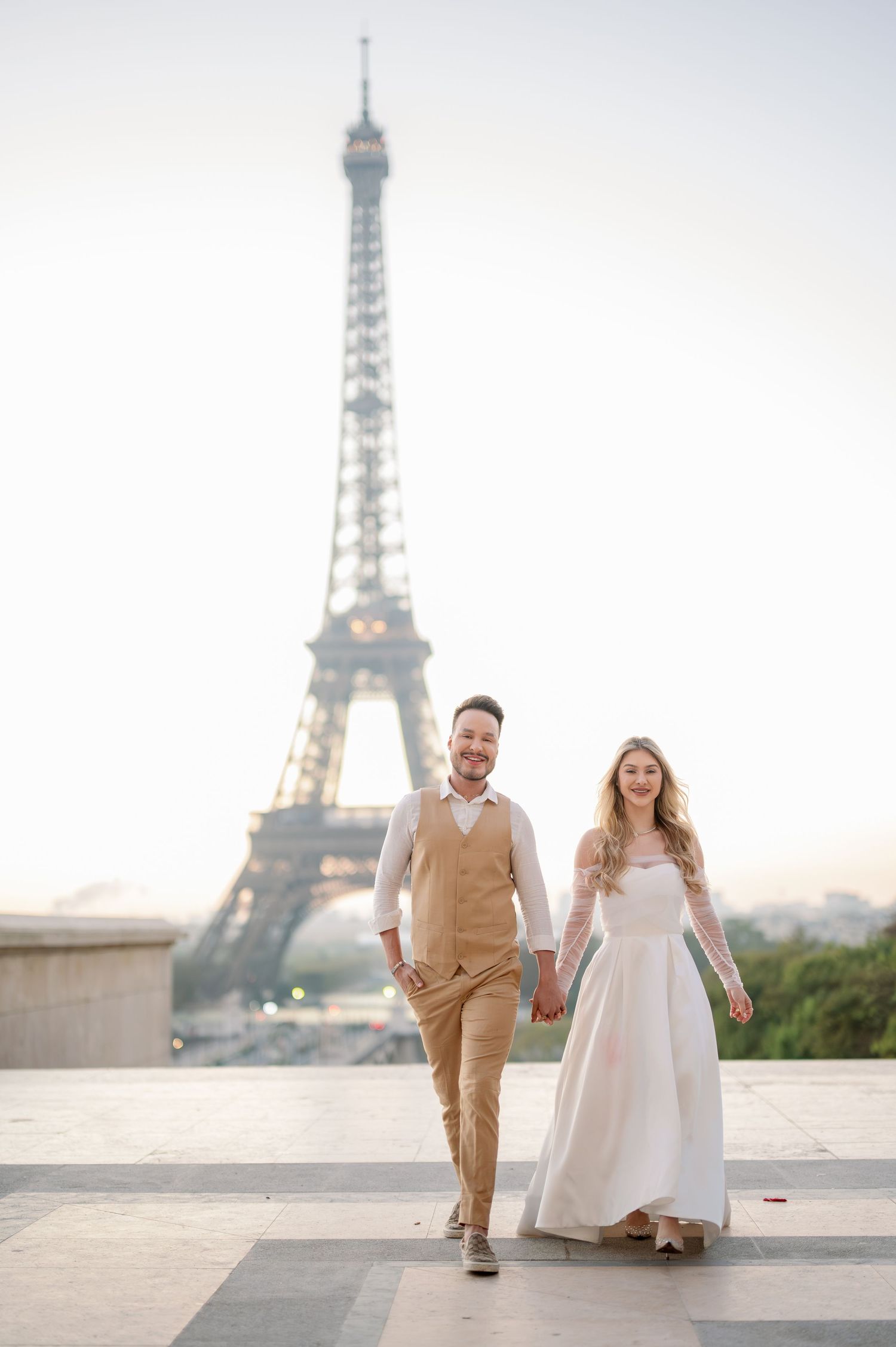 A couple in wedding attire walks hand in hand at sunrise with the Eiffel Tower creating a romantic backdrop in Paris.