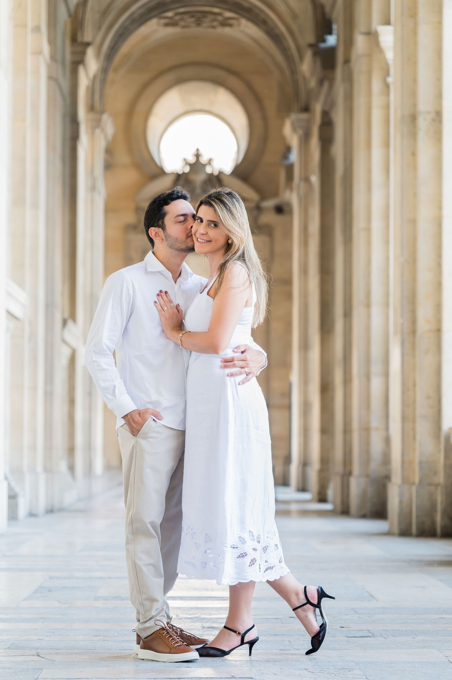 Romantic couple embraces under ornate stone arches in an elegant corridor wearing matching white outfits.