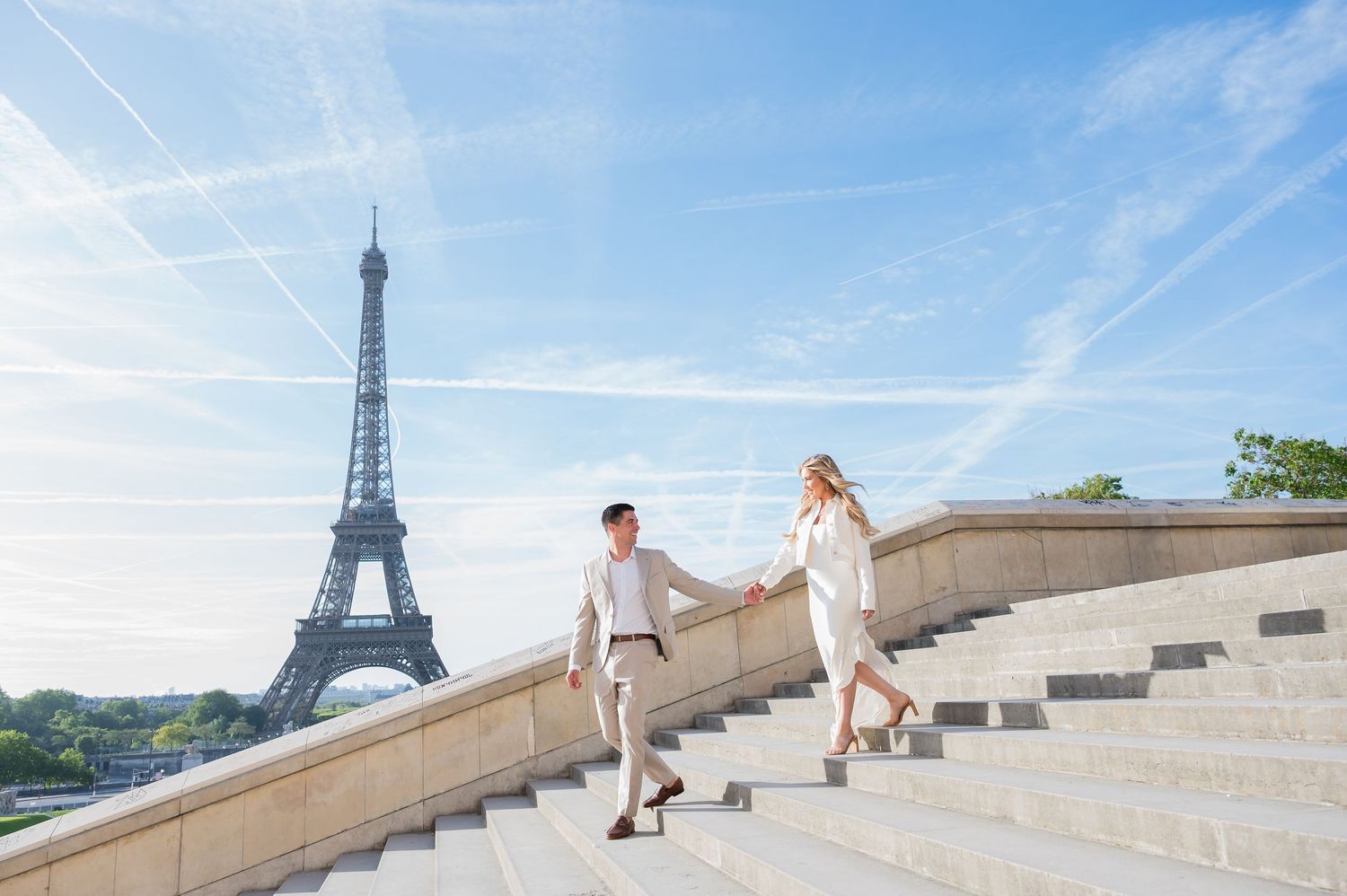 A couple walks down stone steps at Trocadero with the iconic Eiffel Tower in the background on a sunny day in Paris.