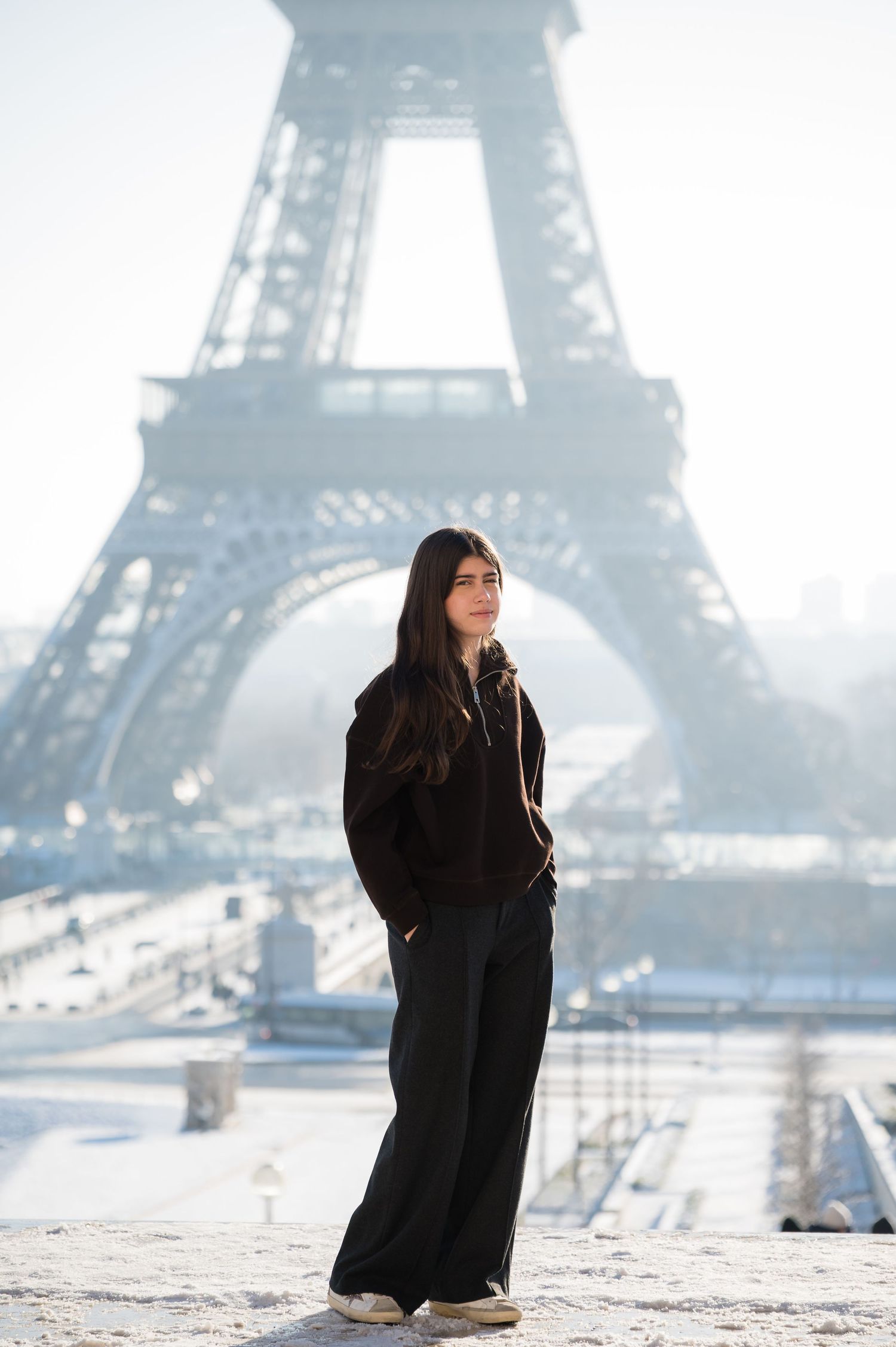 A person in a black coat poses near the Eiffel Tower in Paris on a misty winter morning.
