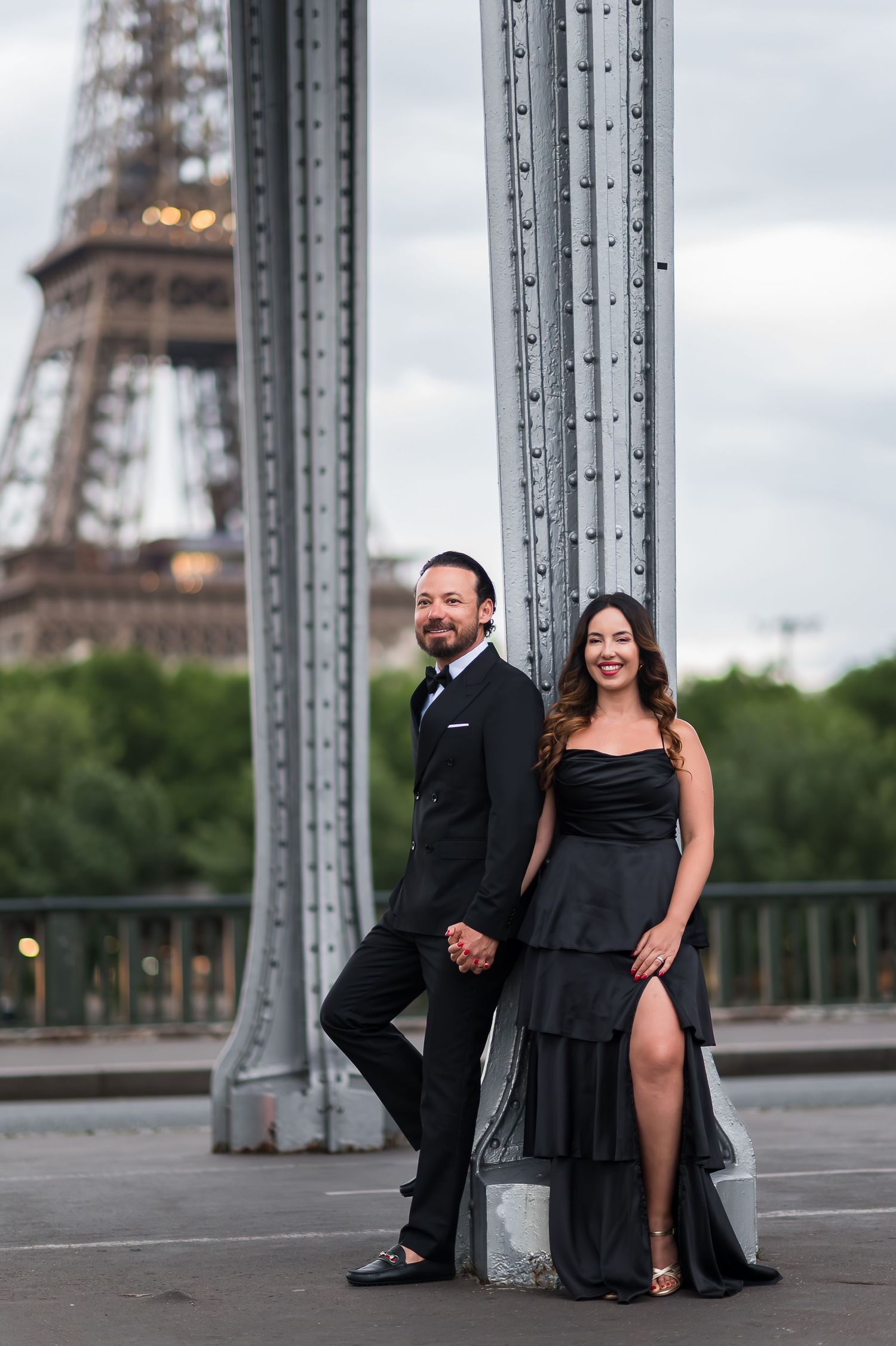 Elegant couple in black formal attire posing on a Paris bridge with the Eiffel Tower in the background.