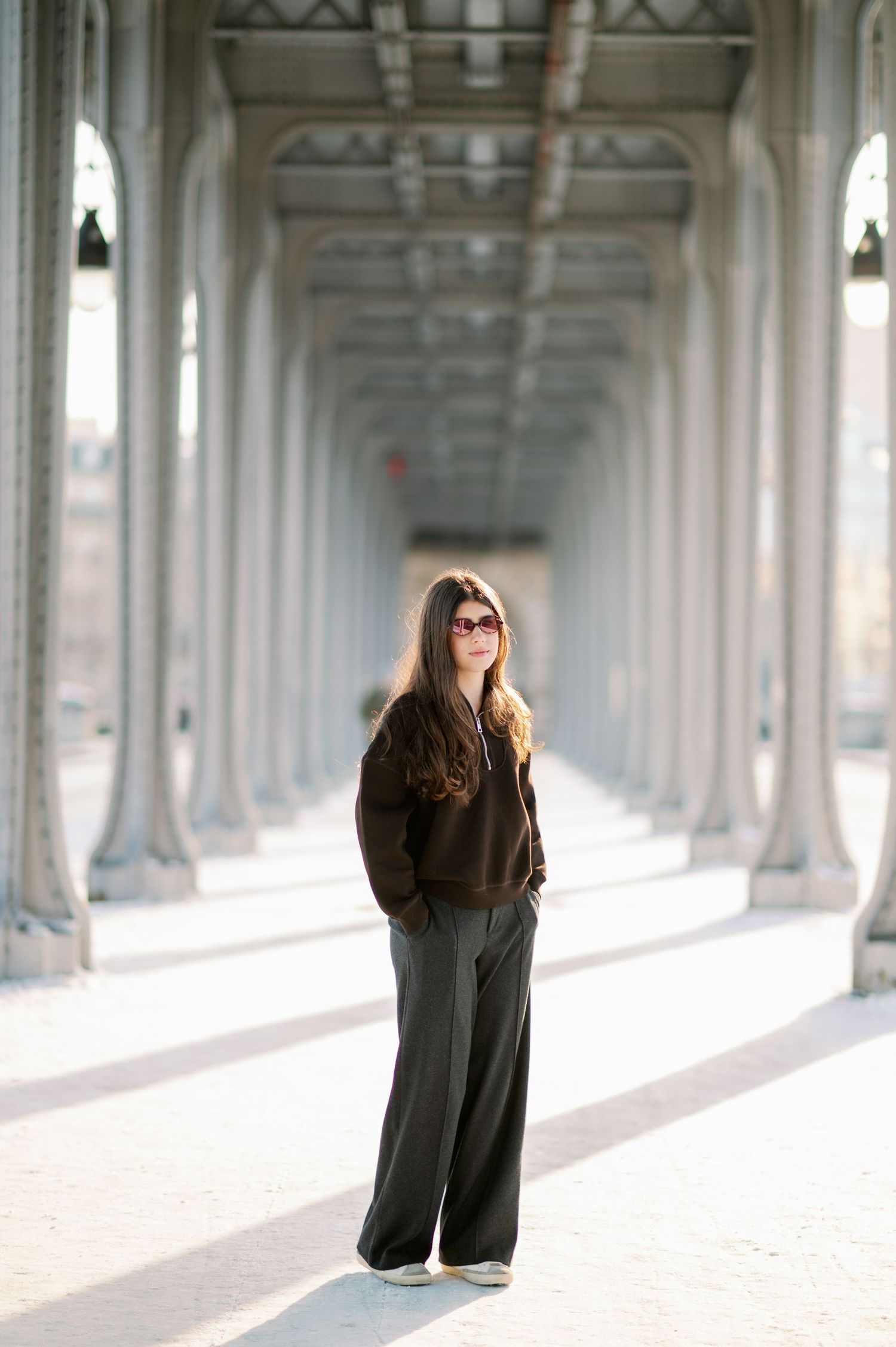 A figure in a black dress stands in a bright colonnade with repeating white arches and columns creating dramatic shadows.