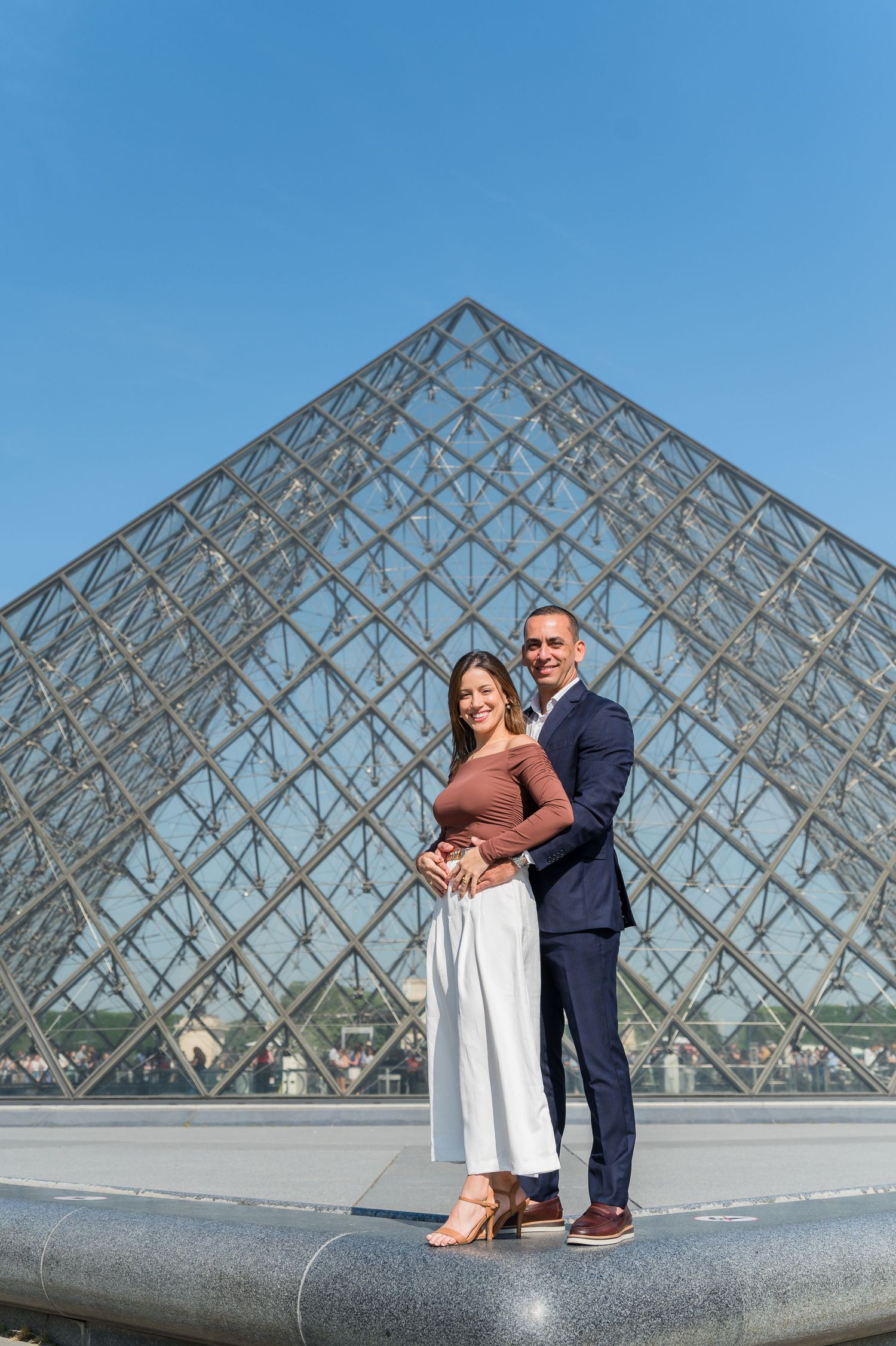 A couple poses together in front of the iconic glass pyramid at the Louvre Museum in Paris on a sunny day.