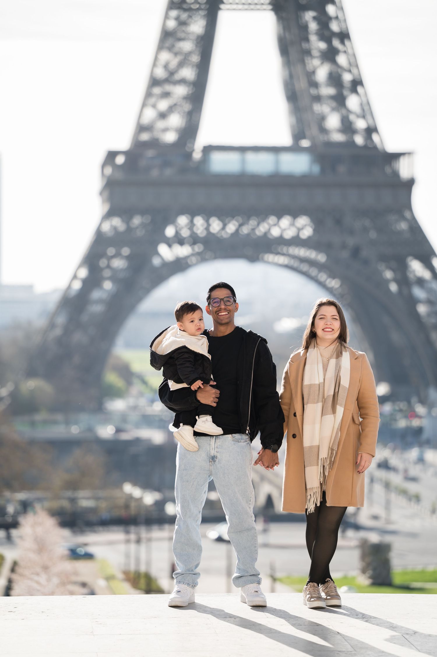 A happy family posing in front of the Eiffel Tower in Paris, with a man holding a baby and a woman beside them.