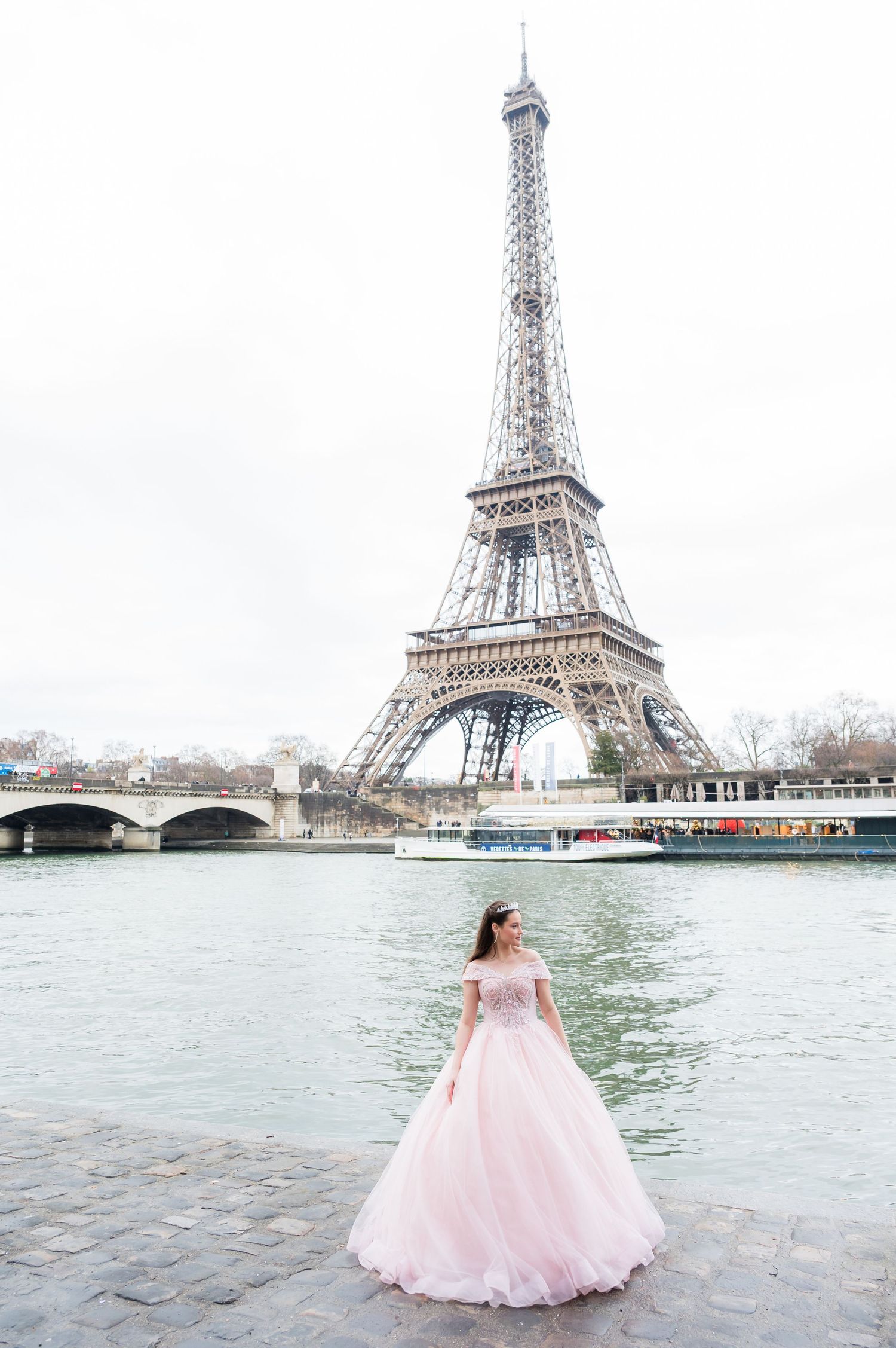 A pink ballgown dress flows gracefully against the backdrop of the Eiffel Tower along the Seine River in Paris.