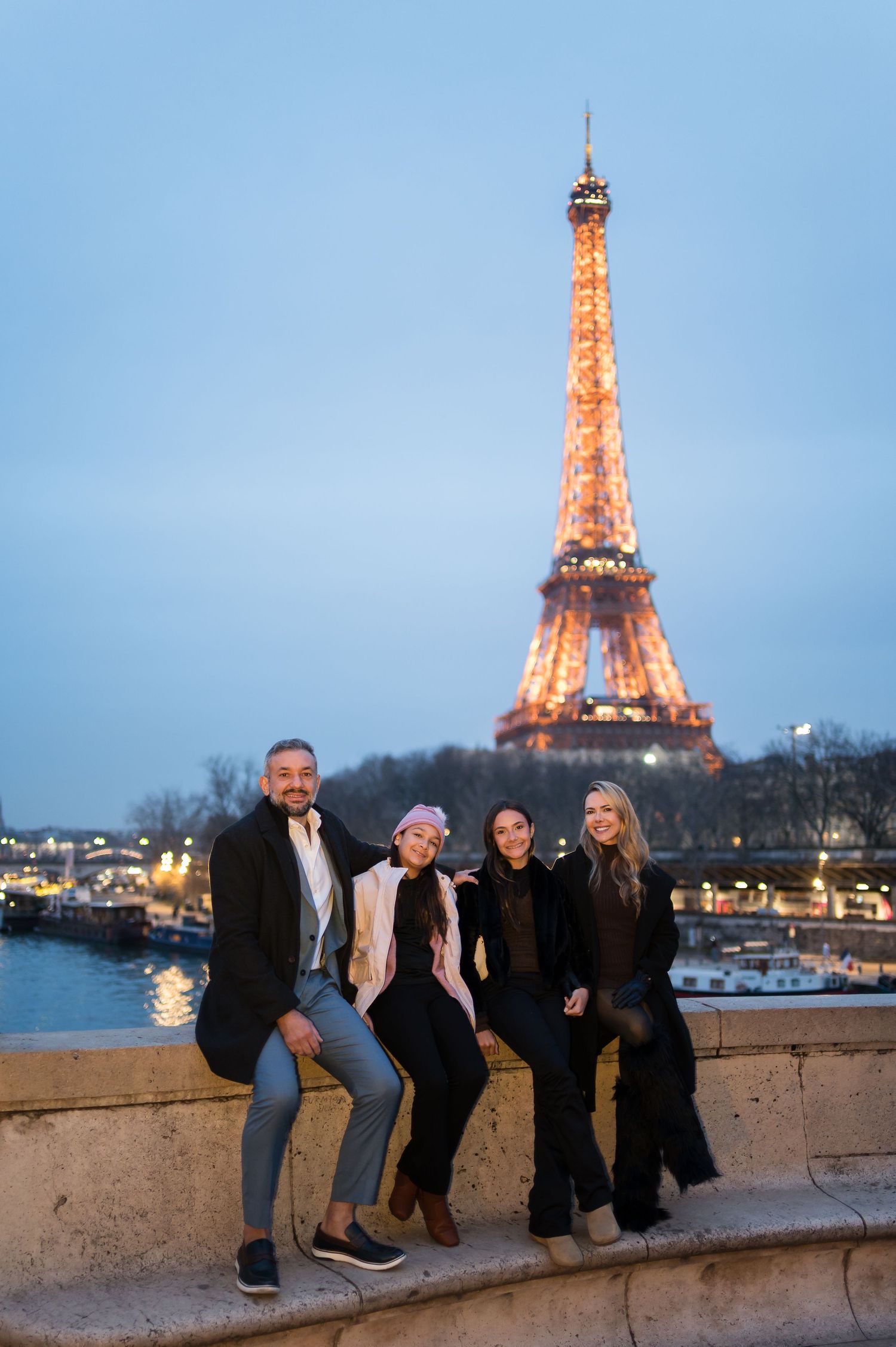 Four people posing on a stone wall with the illuminated Eiffel Tower glowing at dusk in Paris, France.