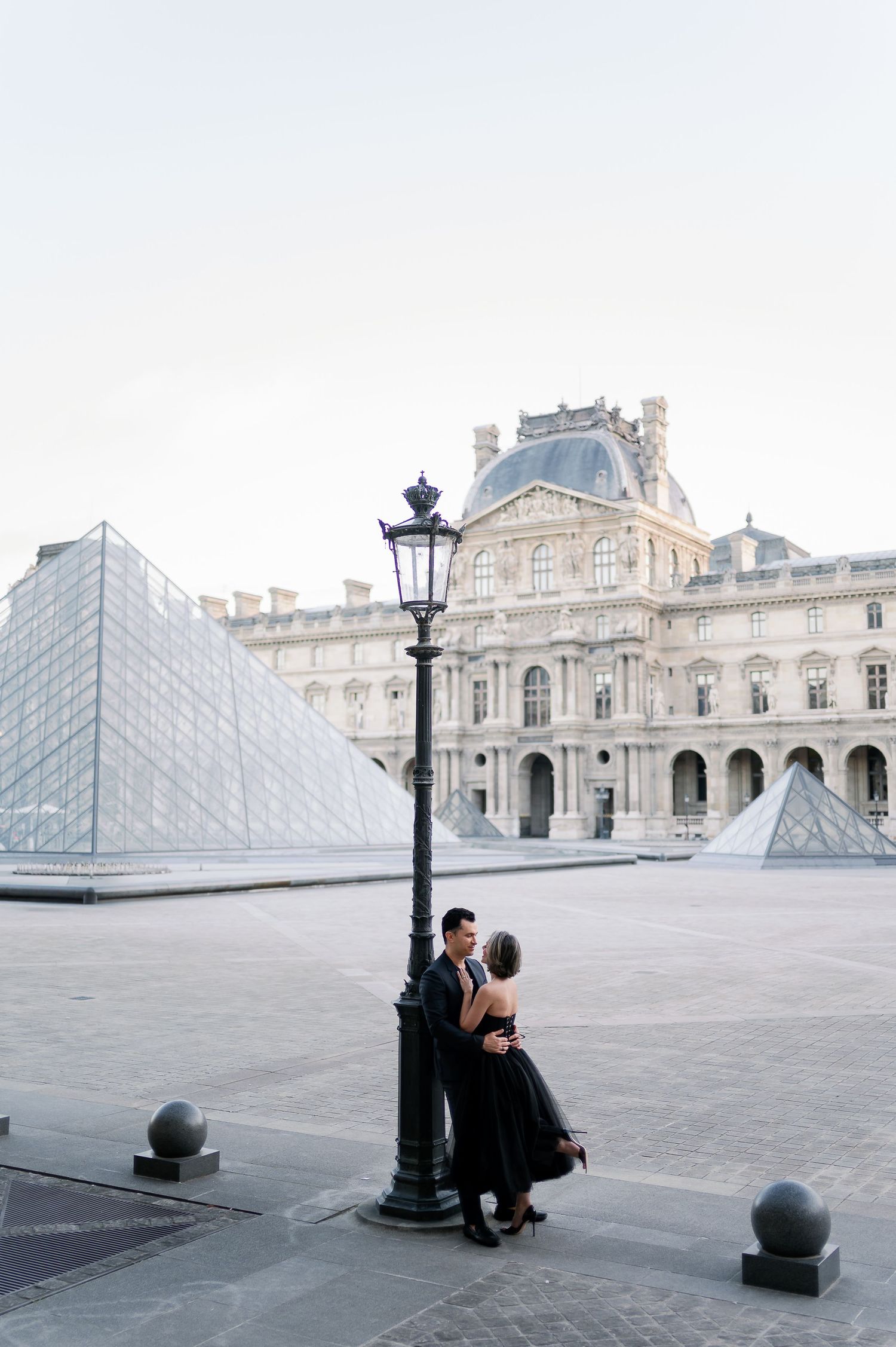 Couple embracing by a lamppost at the Louvre Museum pyramid in Paris, woman in black dress.