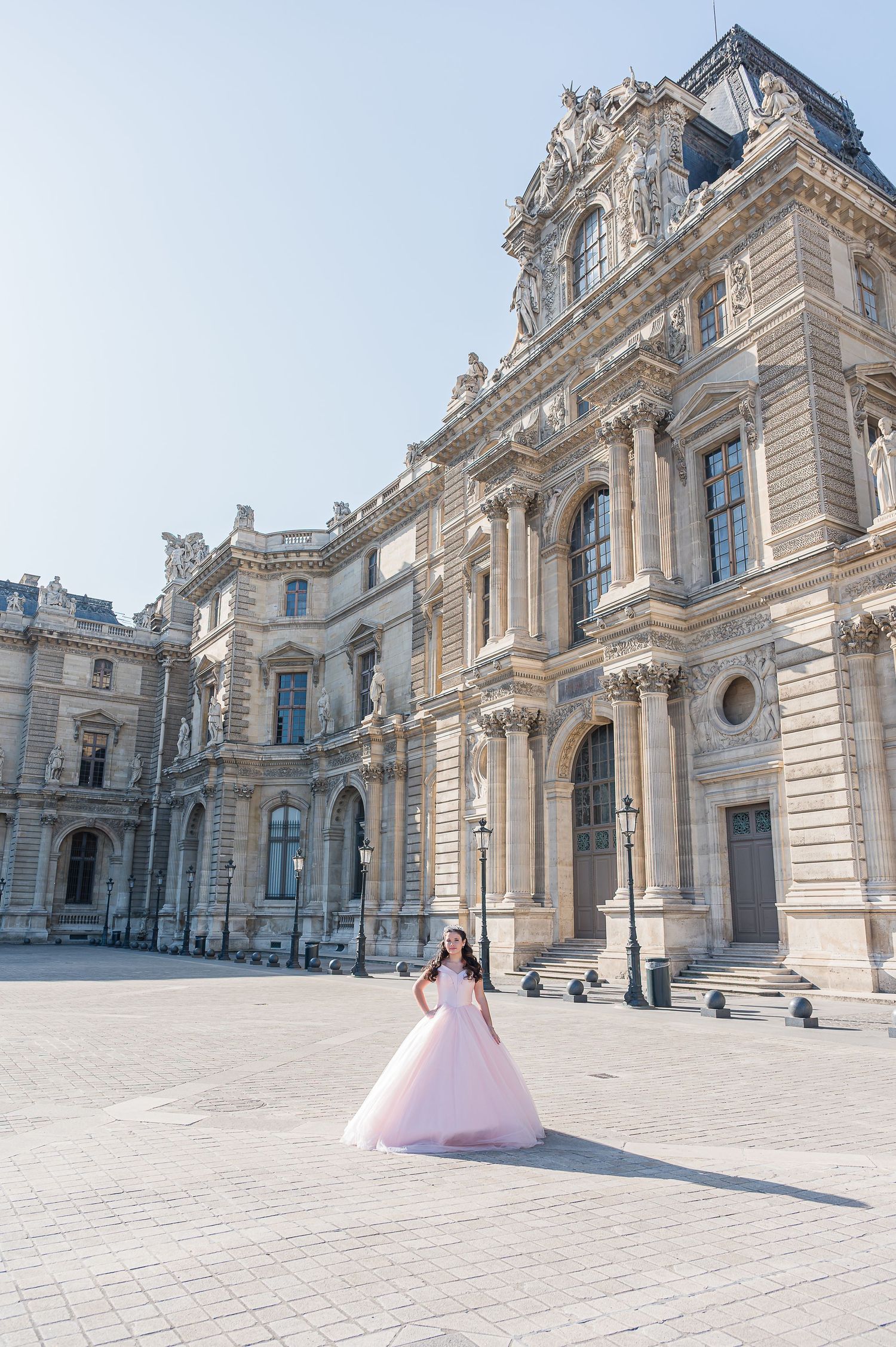 Woman in pink ball gown poses in the courtyard of the Louvre Museum in Paris on a sunny day.