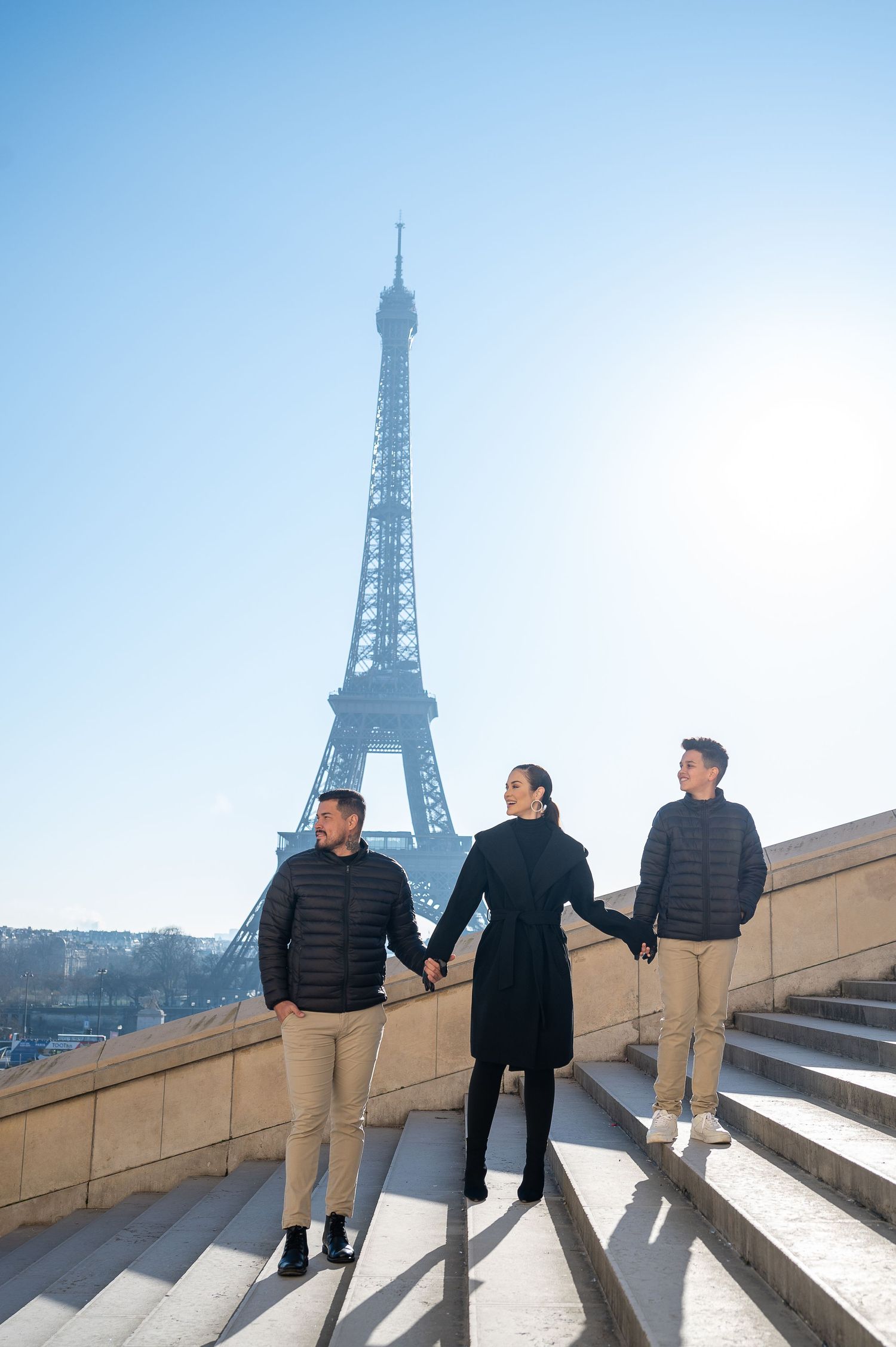 Three people posing on stairs with iconic Eiffel Tower in the background on a sunny day.