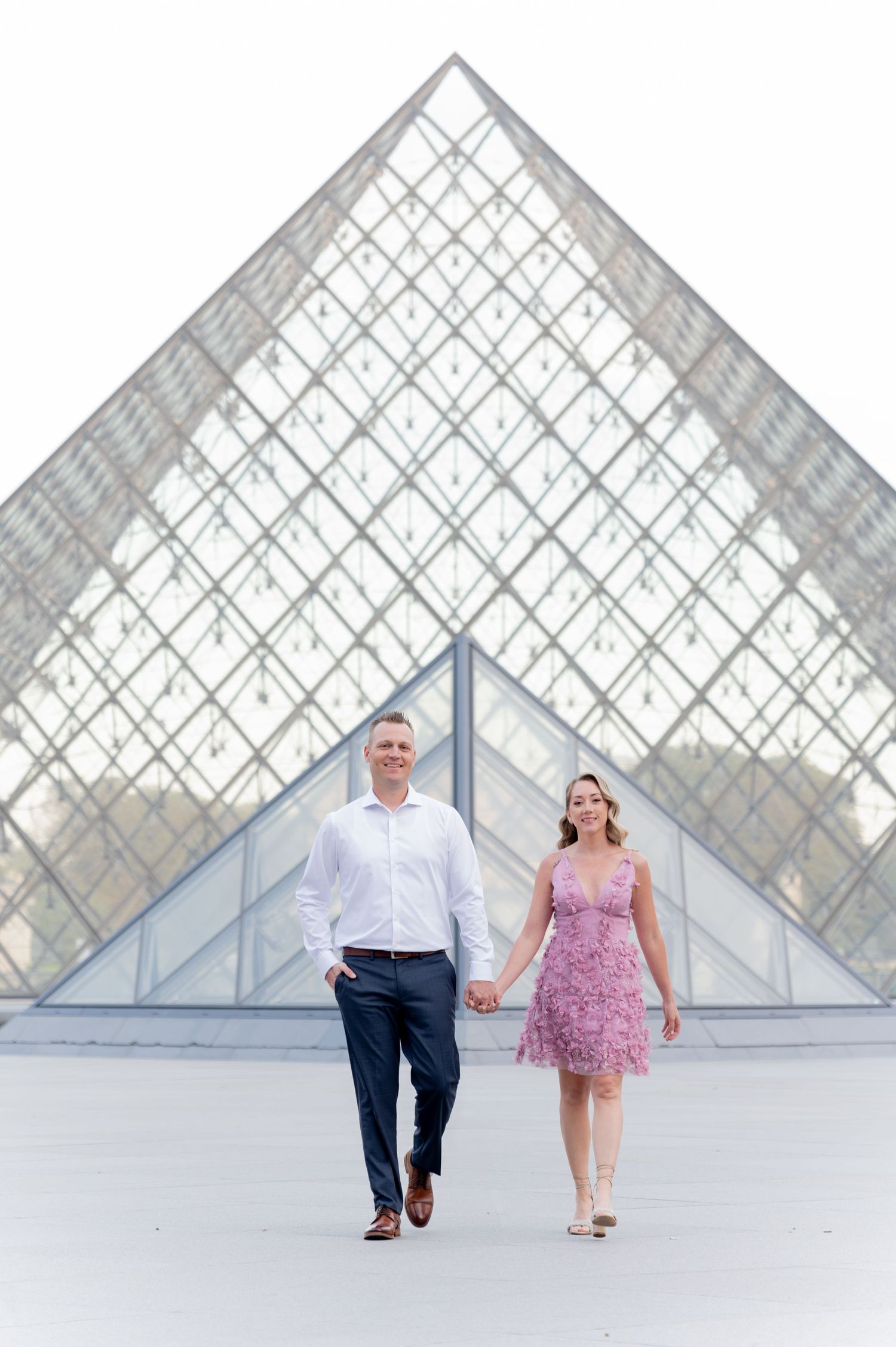 Couple holding hands walking in front of the Louvre Pyramid in Paris, France.