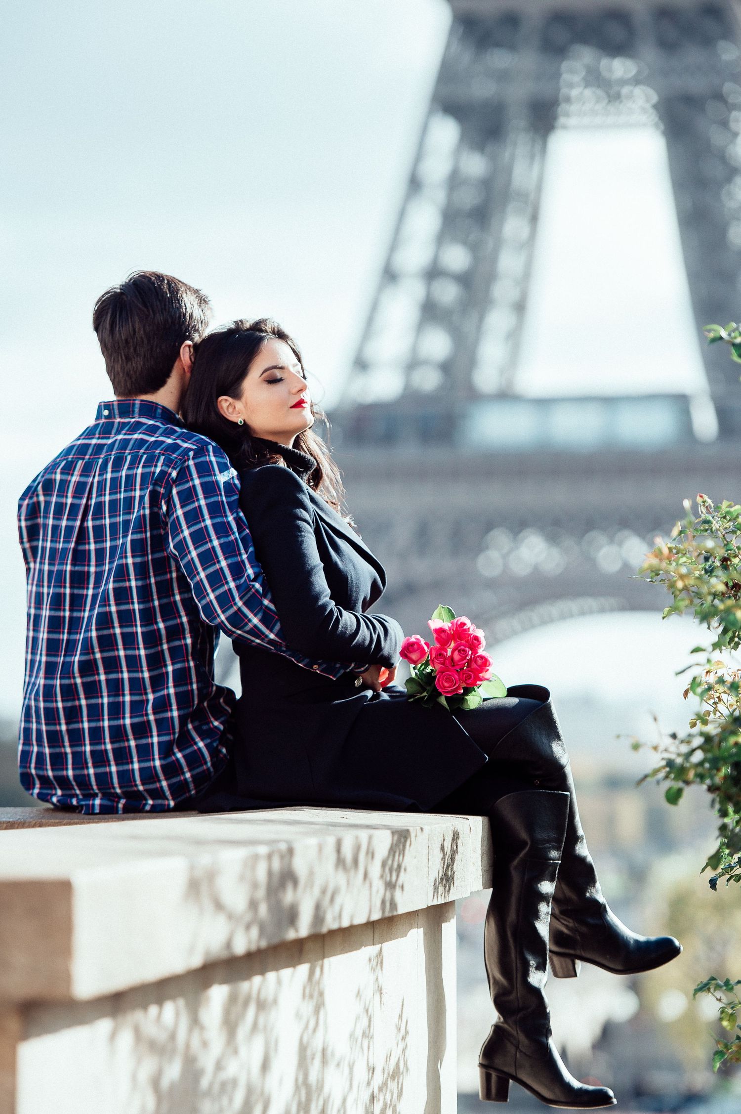 A couple sits together on a ledge with red flowers and the Eiffel Tower in the background in Paris.