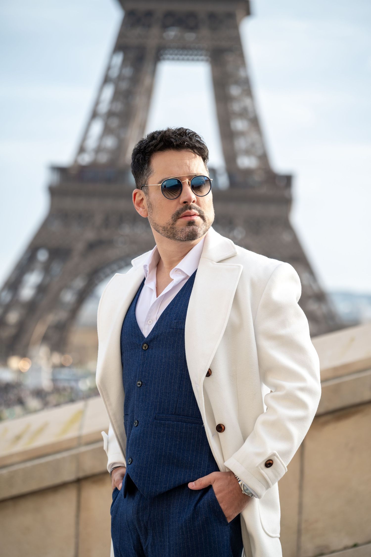 Stylish man in cream blazer and navy vest poses confidently against Eiffel Tower backdrop in Paris.