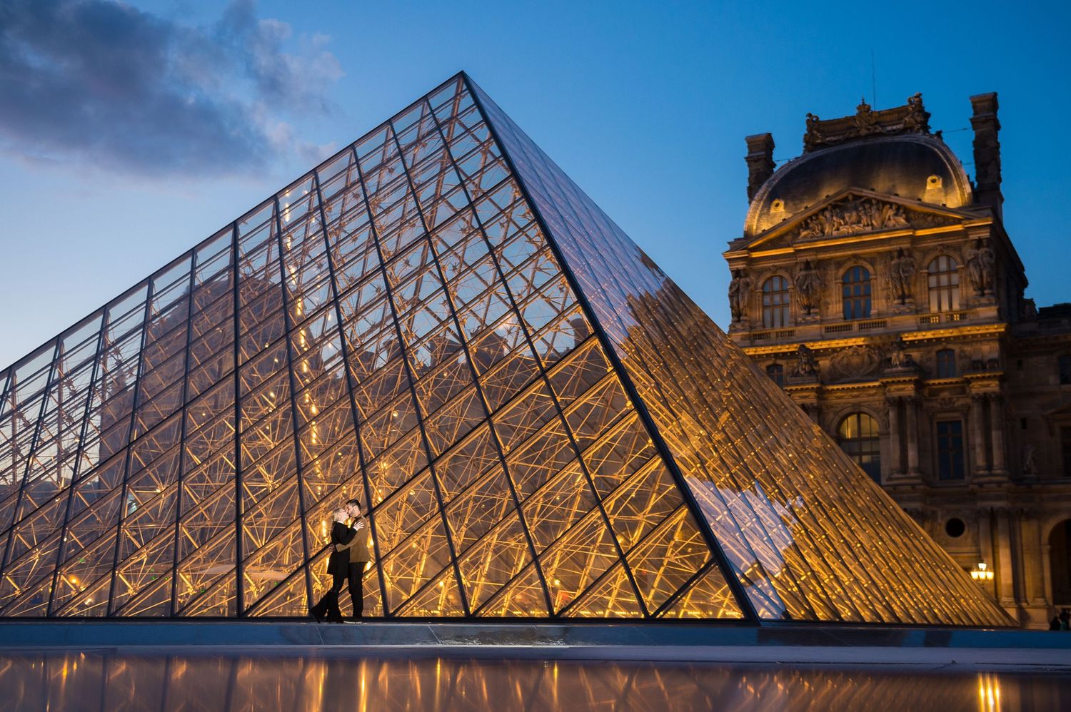 Illuminated glass pyramid entrance of the Louvre Museum in Paris glows against historic palace architecture at dusk.