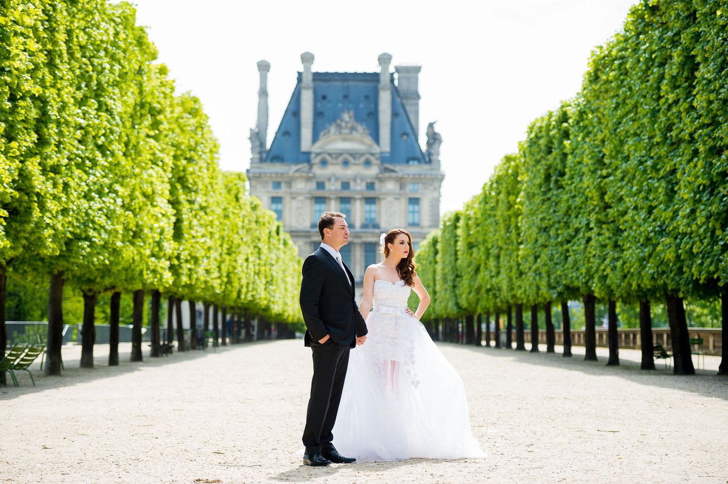 A couple poses in formal attire along a tree-lined path with the Louvre Palace in Paris visible in the background.