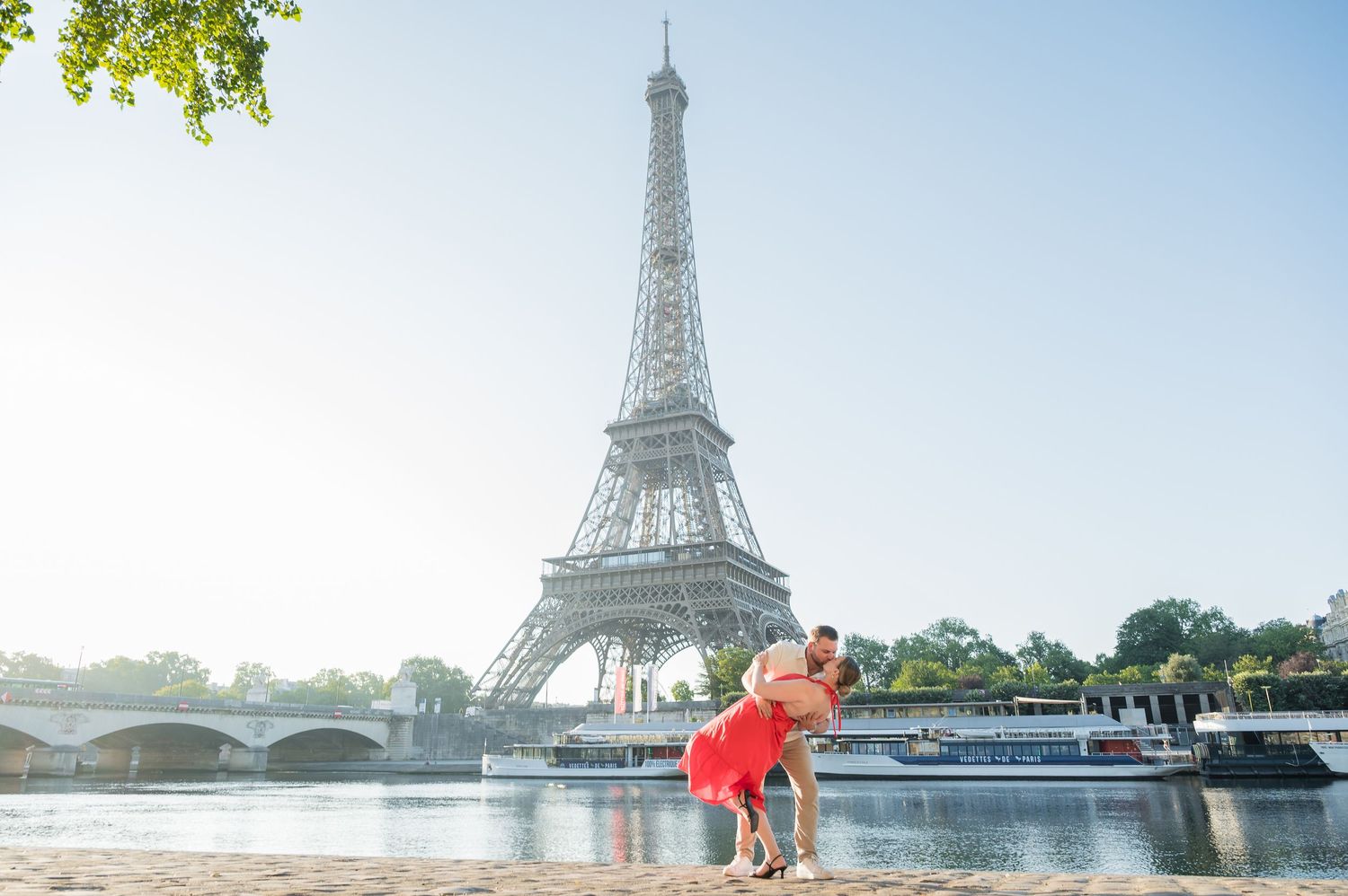 Couple stands together near the Eiffel Tower in Paris with buildings and green trees in the background.