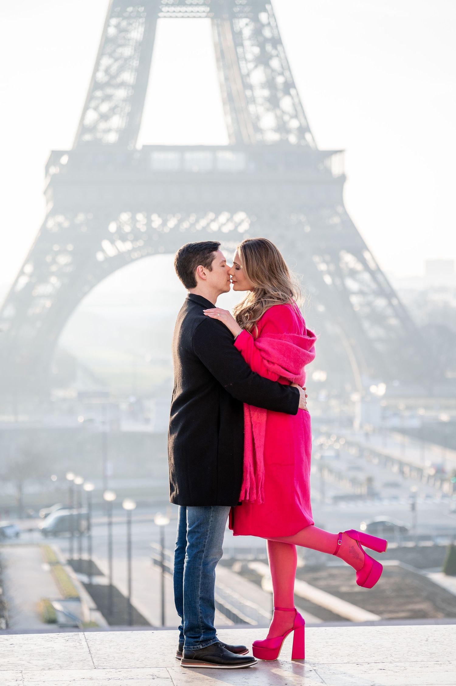 Romantic kiss scene in front of misty Eiffel Tower with bright red coat adding vibrant pop of color.
