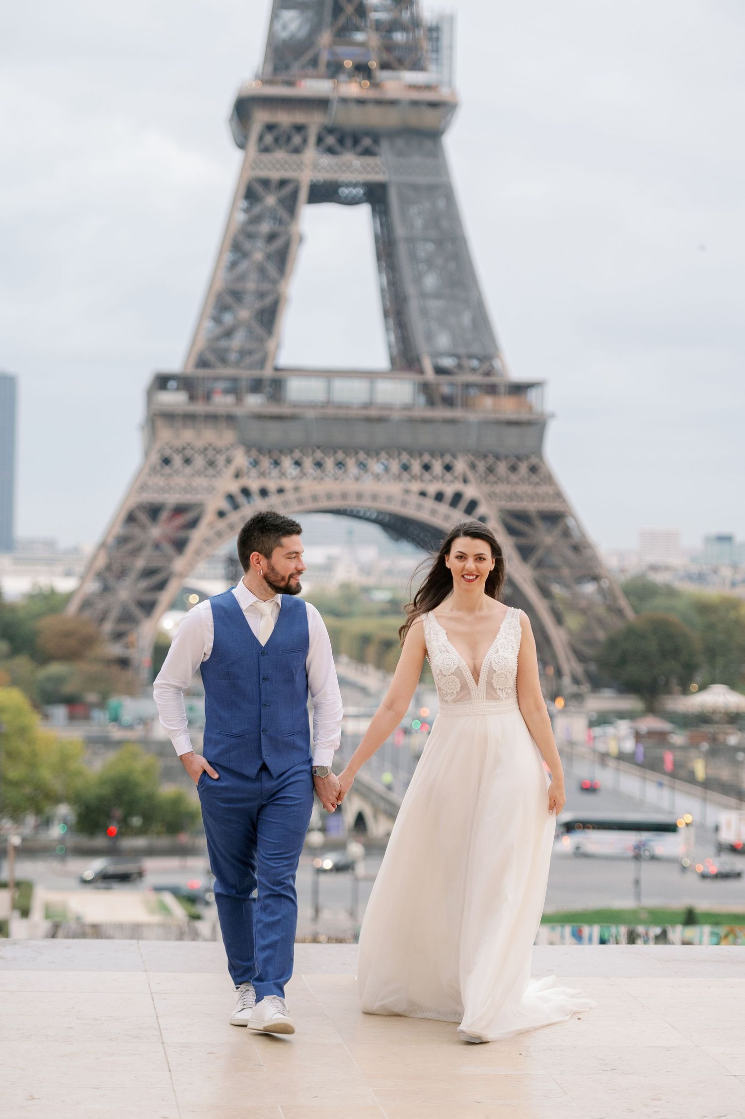 A couple in wedding attire walks hand in hand at Trocadero with multiple views of the Eiffel Tower in Paris, France.