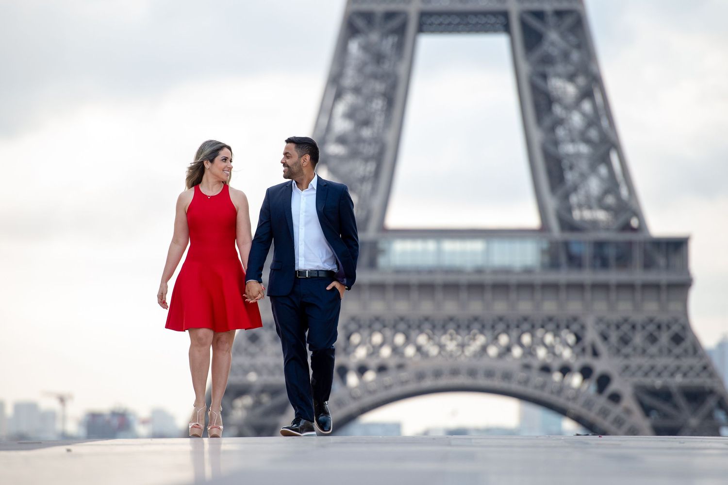 A romantic couple in red dress and suit poses in front of the iconic Eiffel Tower in Paris on a cloudy day.