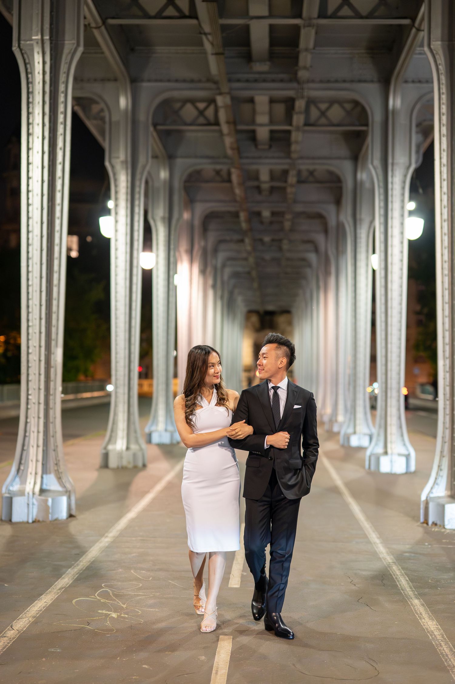 Couple walking together under illuminated bridge architecture with metal columns at dusk.