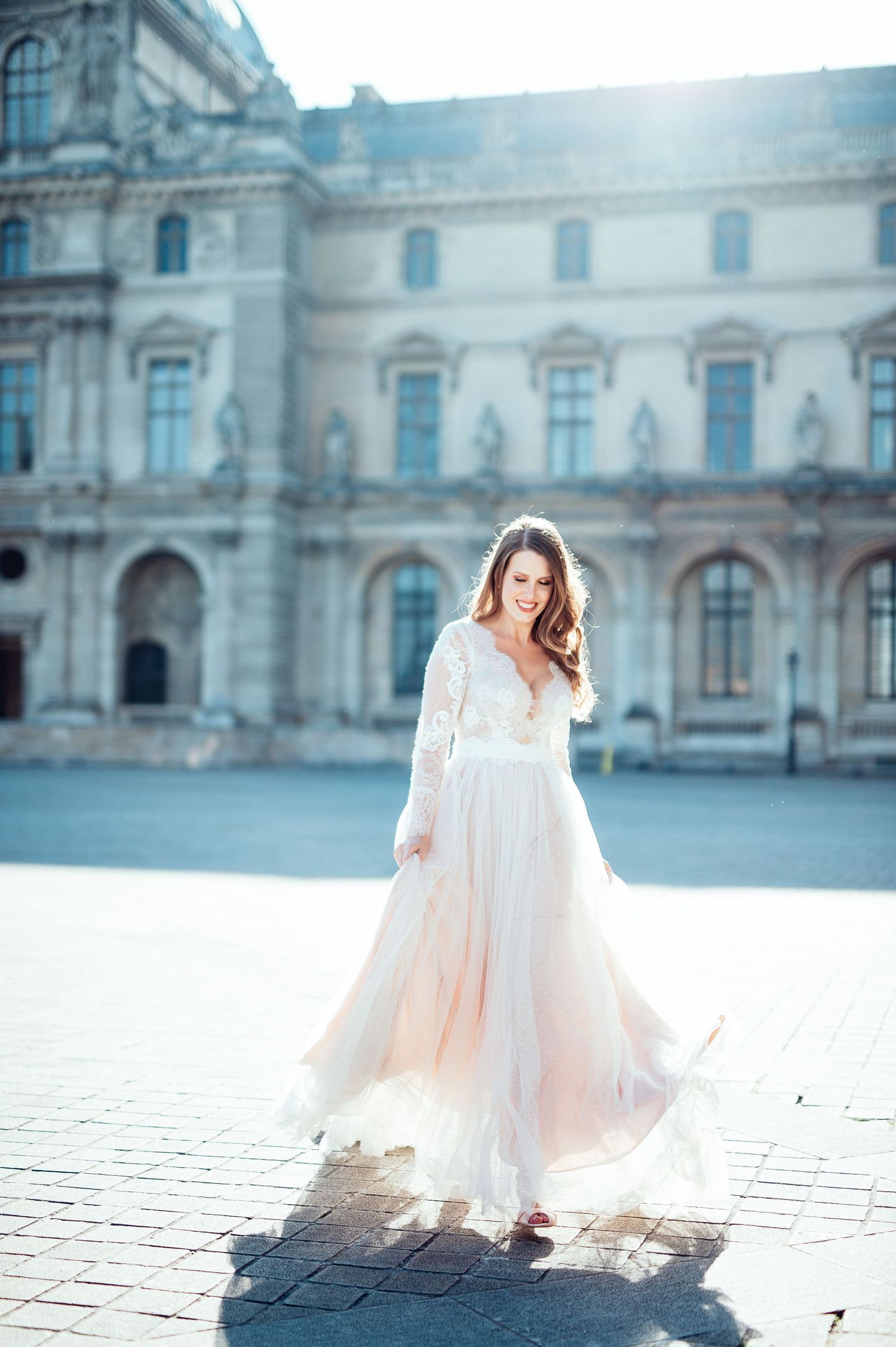 Woman in flowing white gown twirls in front of ornate European palace building in soft sunlight.