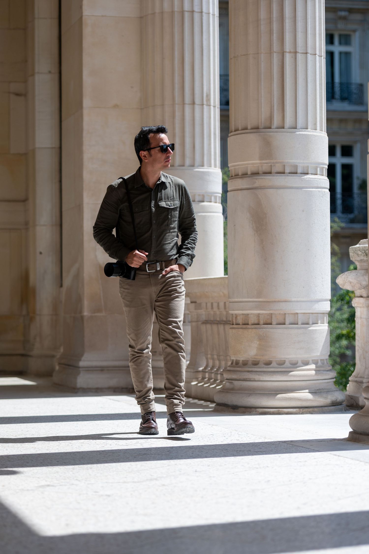 Someone in sunglasses, a green jacket and gray pants leans casually against classic stone columns in soft natural light.