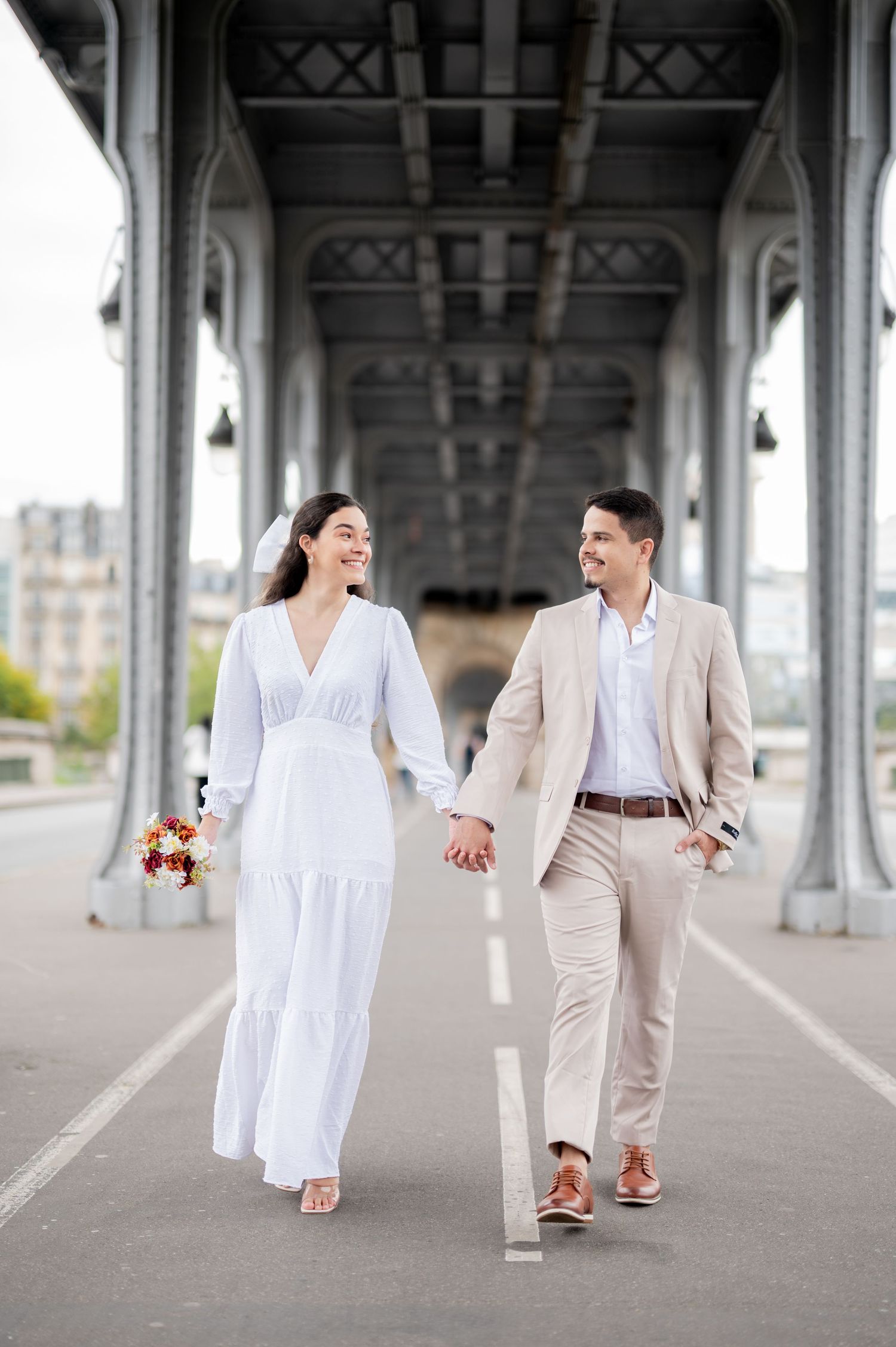A couple in coordinated white and beige outfits walking hand in hand under an architectural bridge walkway.