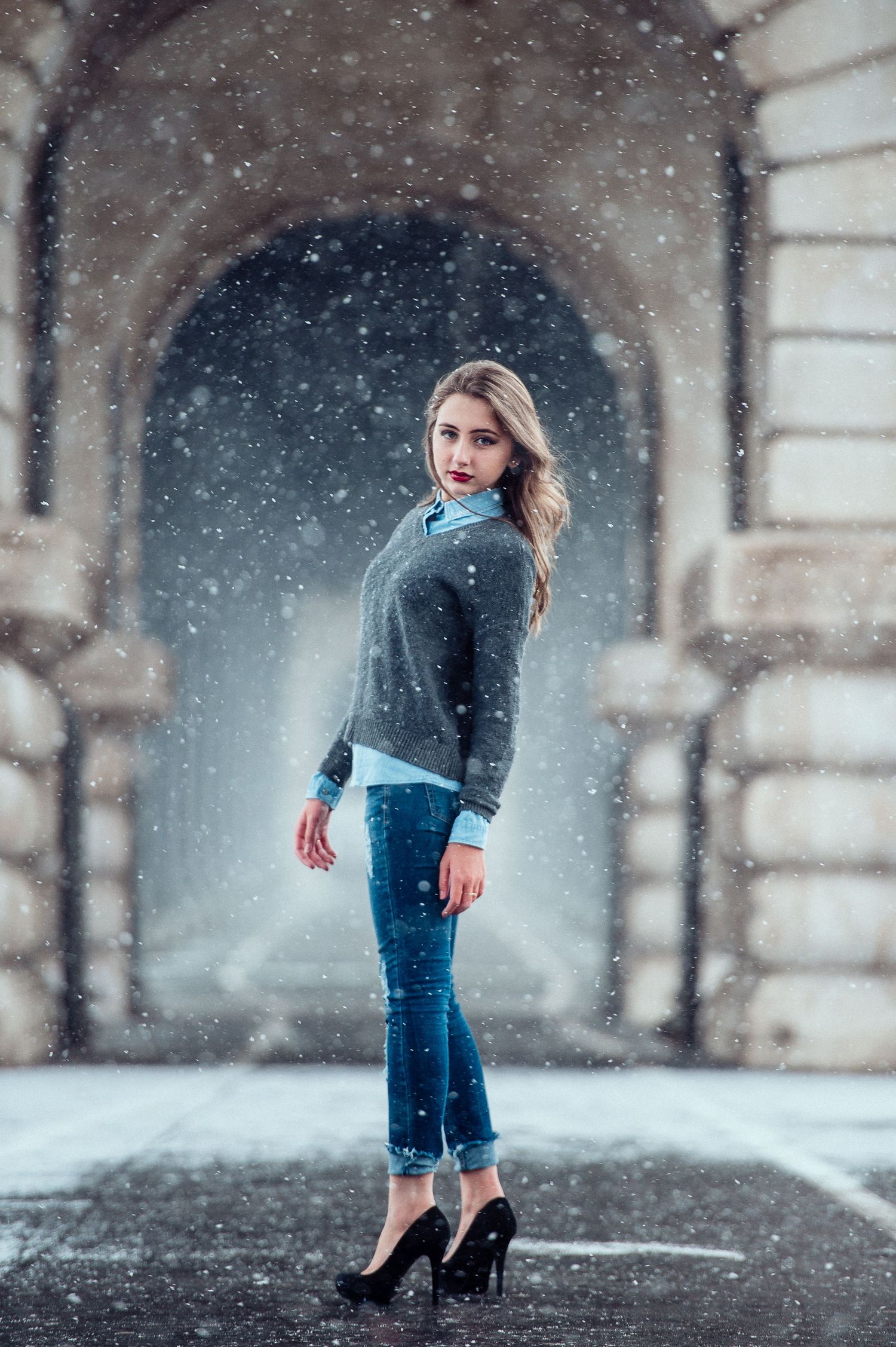 A person in a gray sweater and jeans poses in an archway while snow falls in a romantic winter scene.