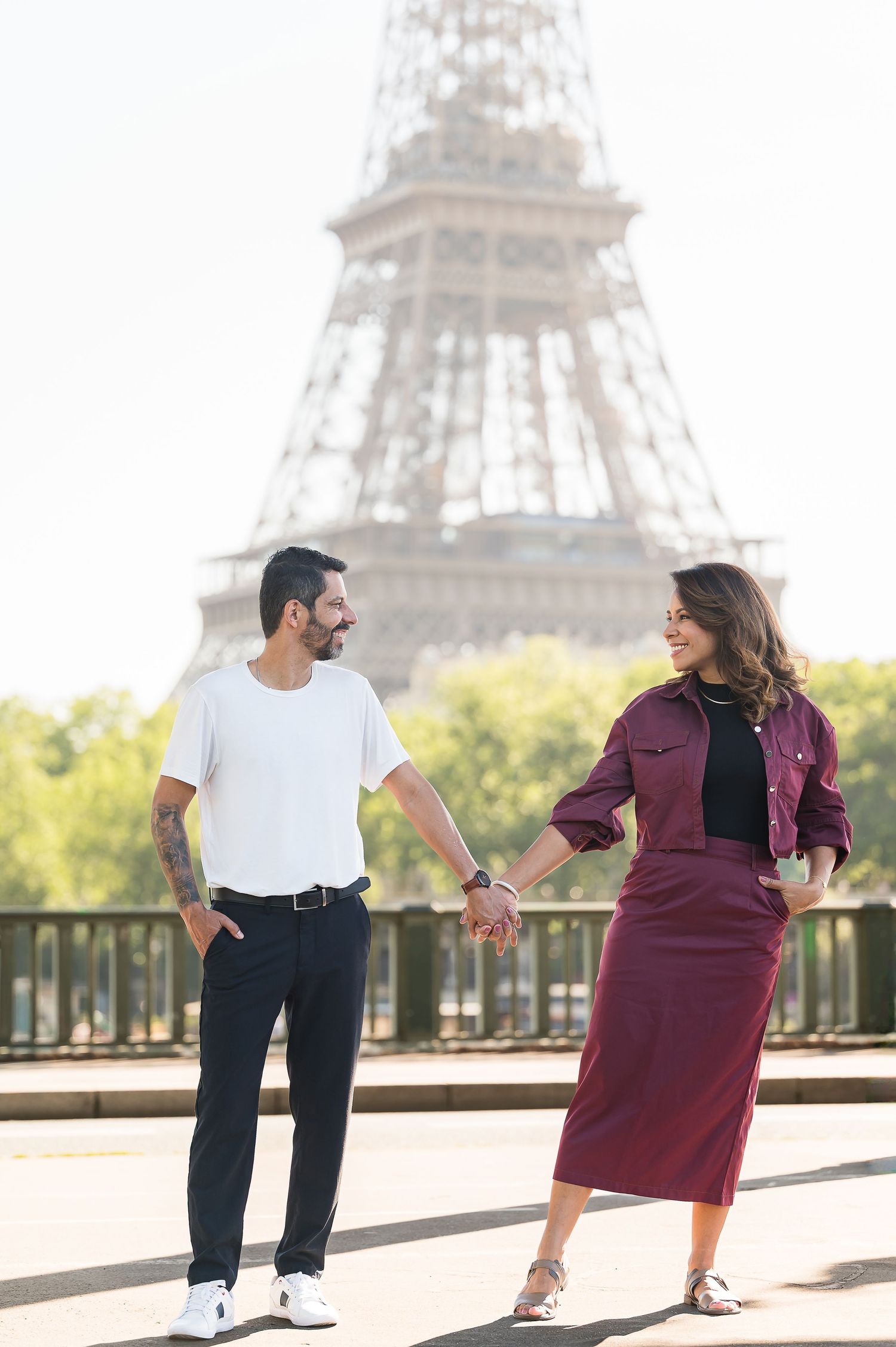 Couple holding hands in front of the Eiffel Tower in Paris, France, smiling at each other on a sunny day.