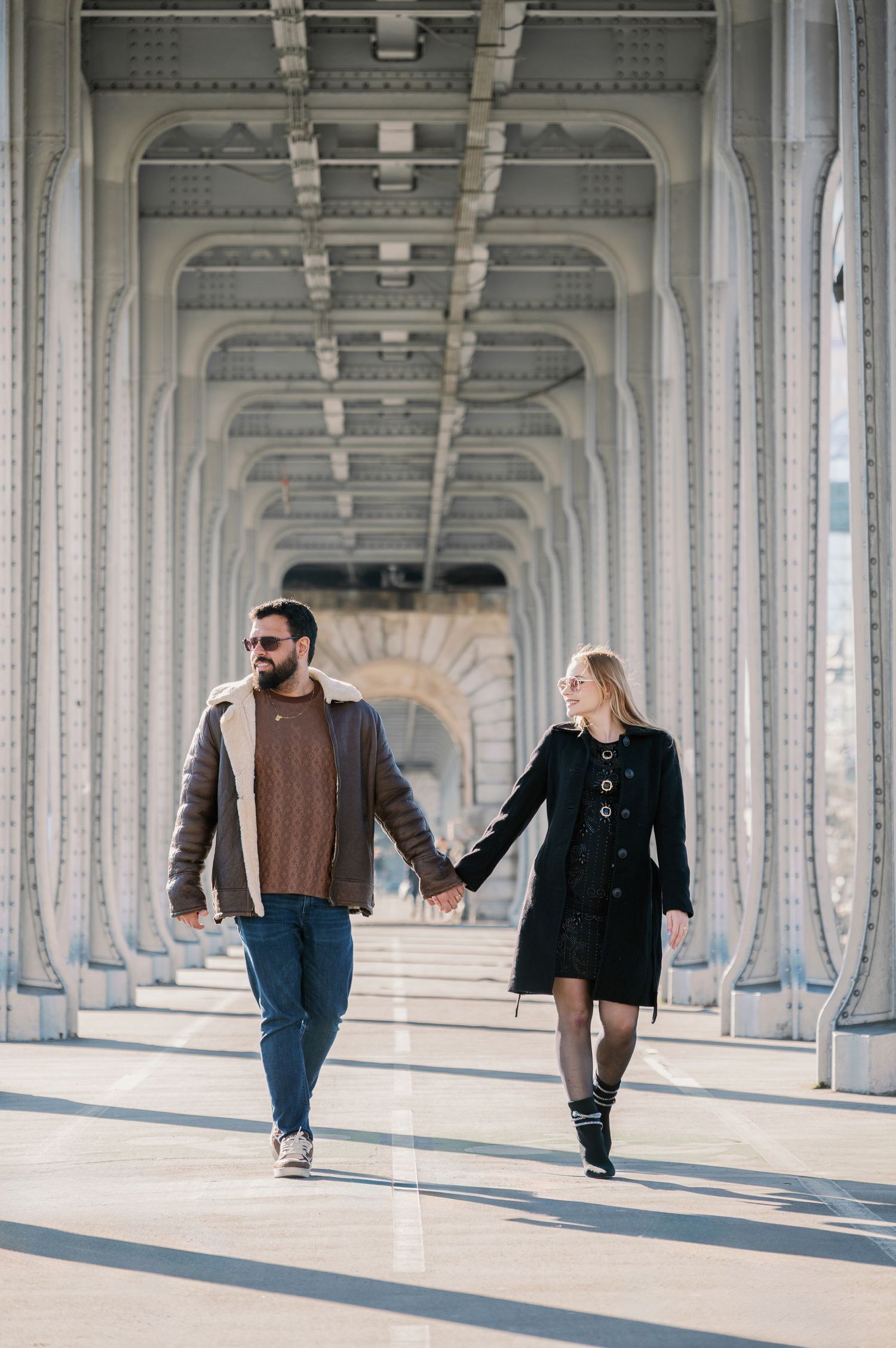 A couple strolls hand in hand under the geometric architecture of a Parisian bridge.