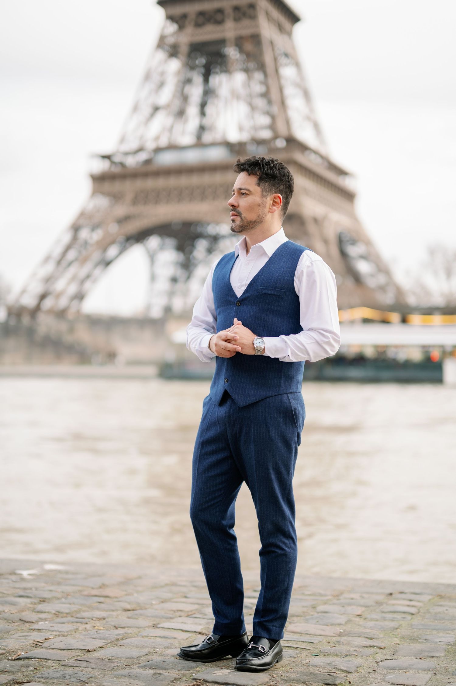 Fashion portrait of well-dressed man in navy suit and white shirt standing by Eiffel Tower in Paris.