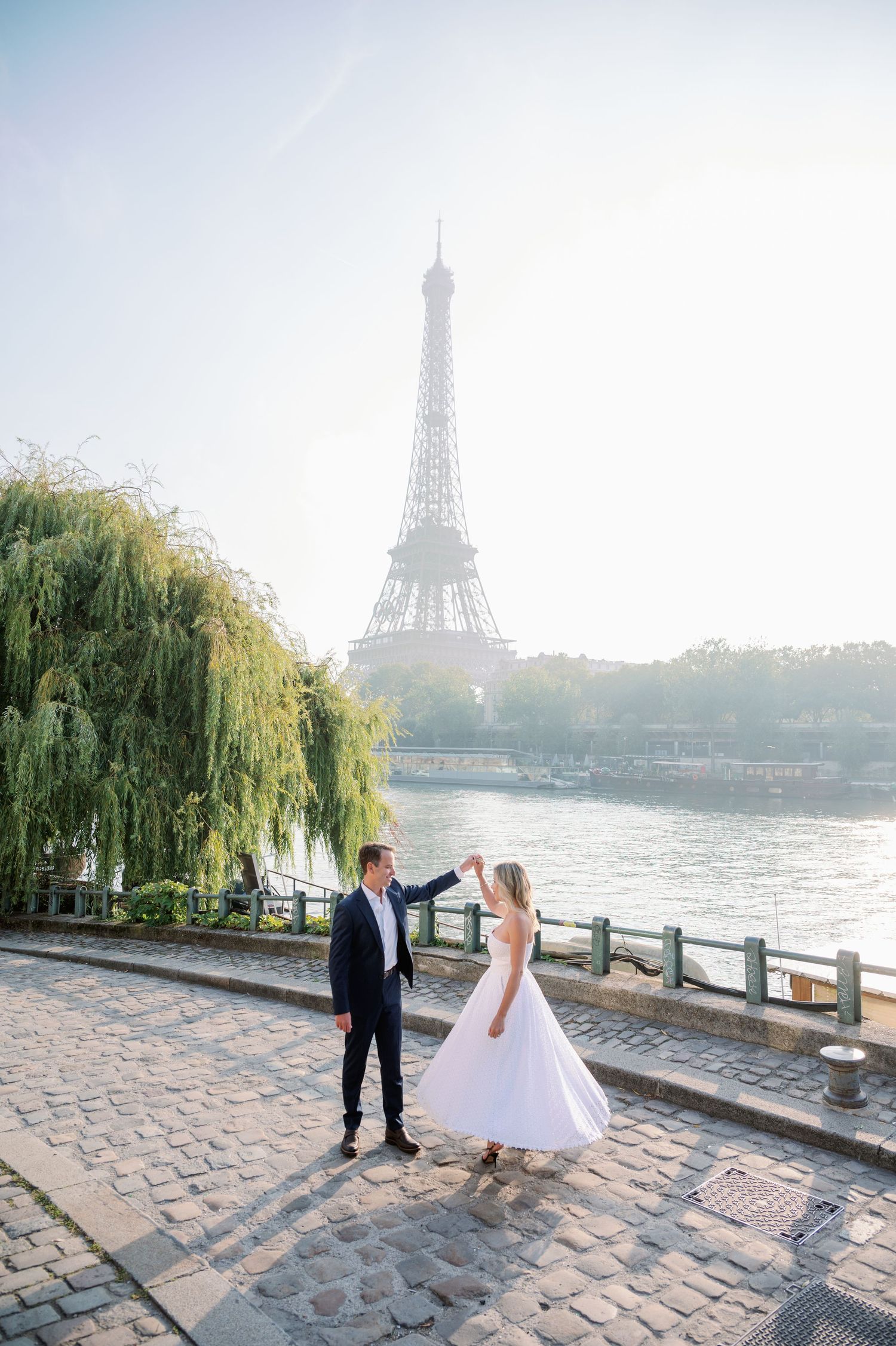 A romantic wedding couple dances along the cobblestone path by the Seine River with the Eiffel Tower in soft morning light.