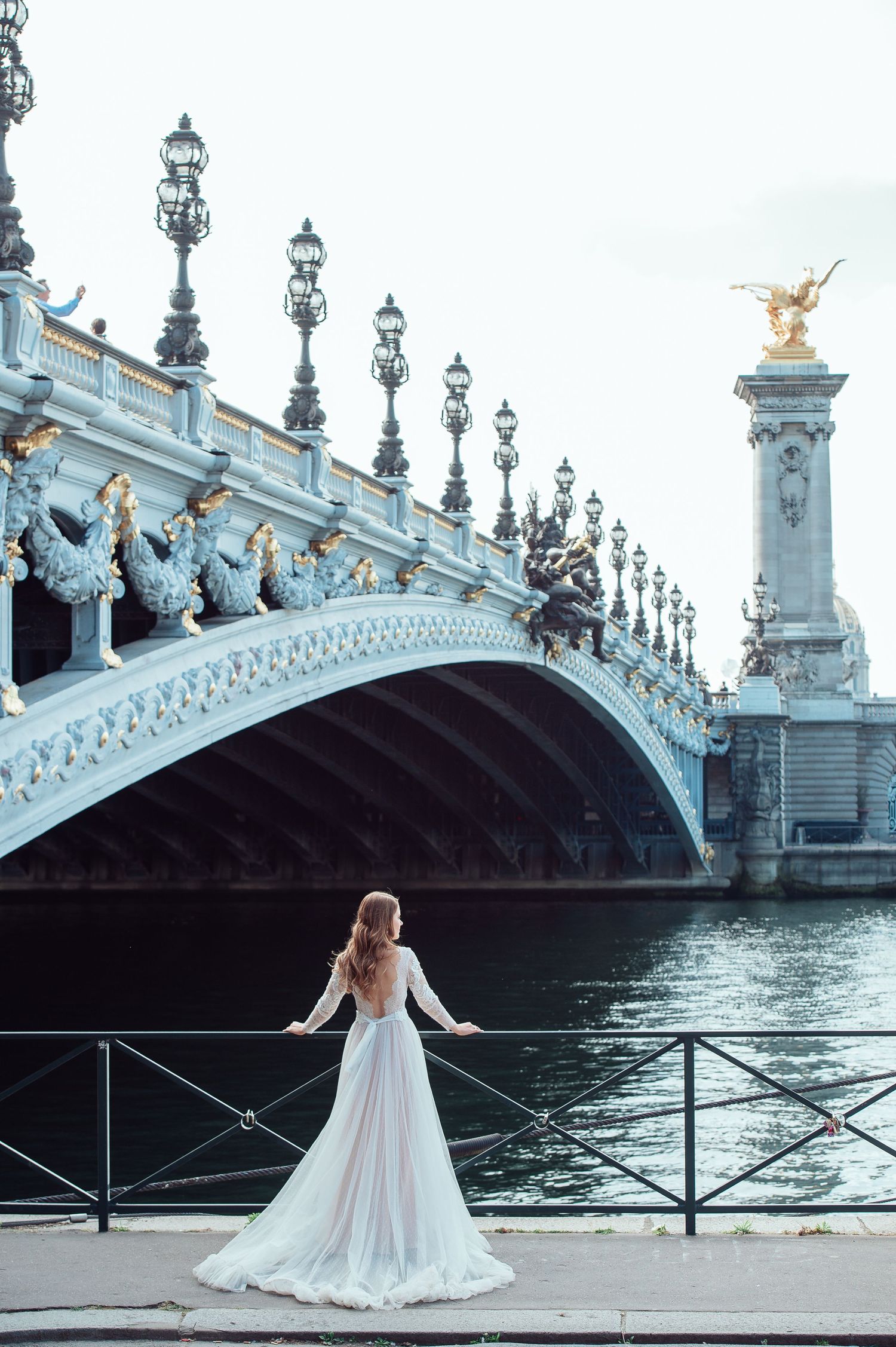 Woman in white wedding dress poses on bridge near Alexandre III bridge and Seine River in Paris.
