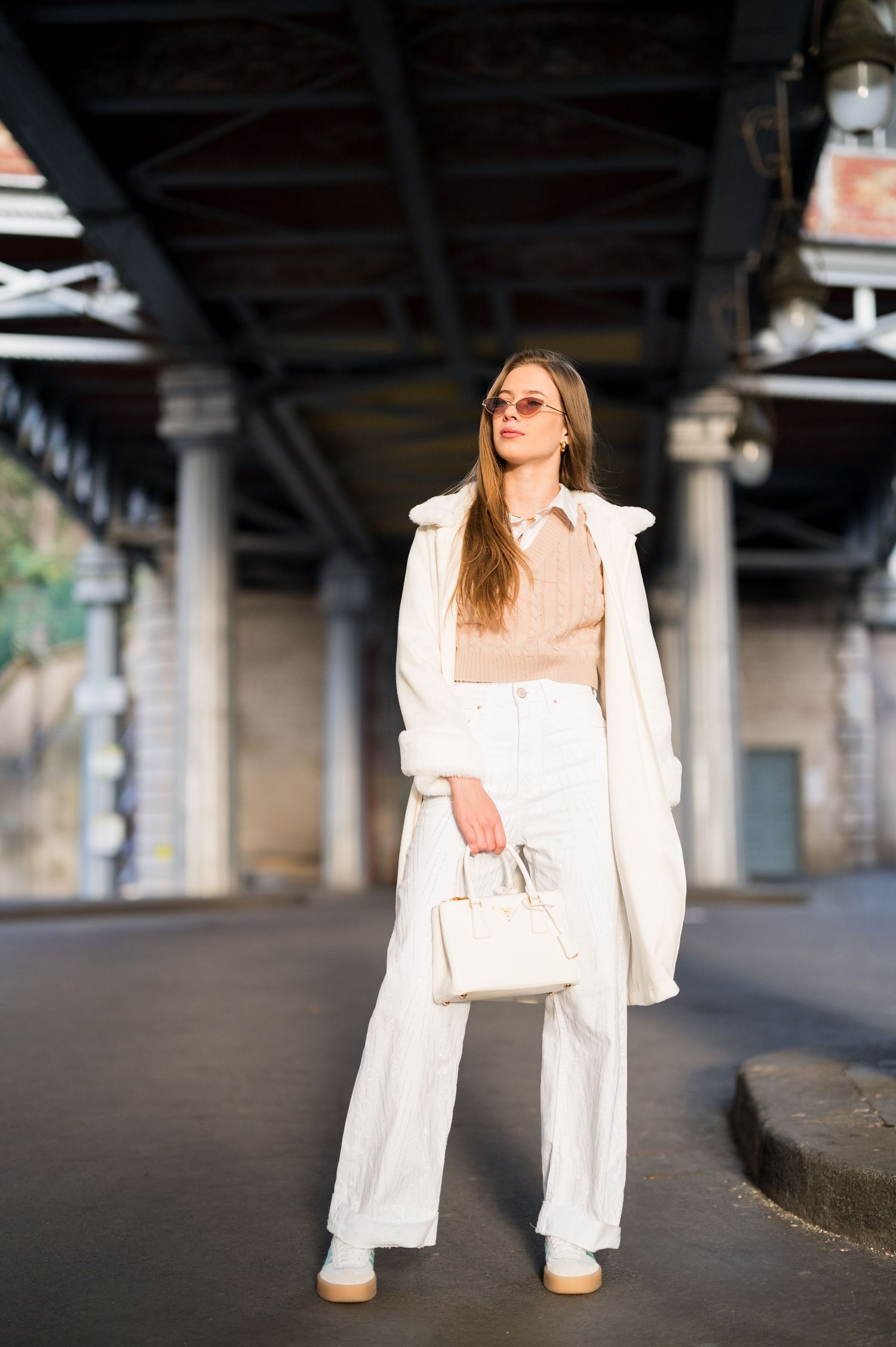 Person in all-white ensemble with handbag posing beneath urban bridge architecture.