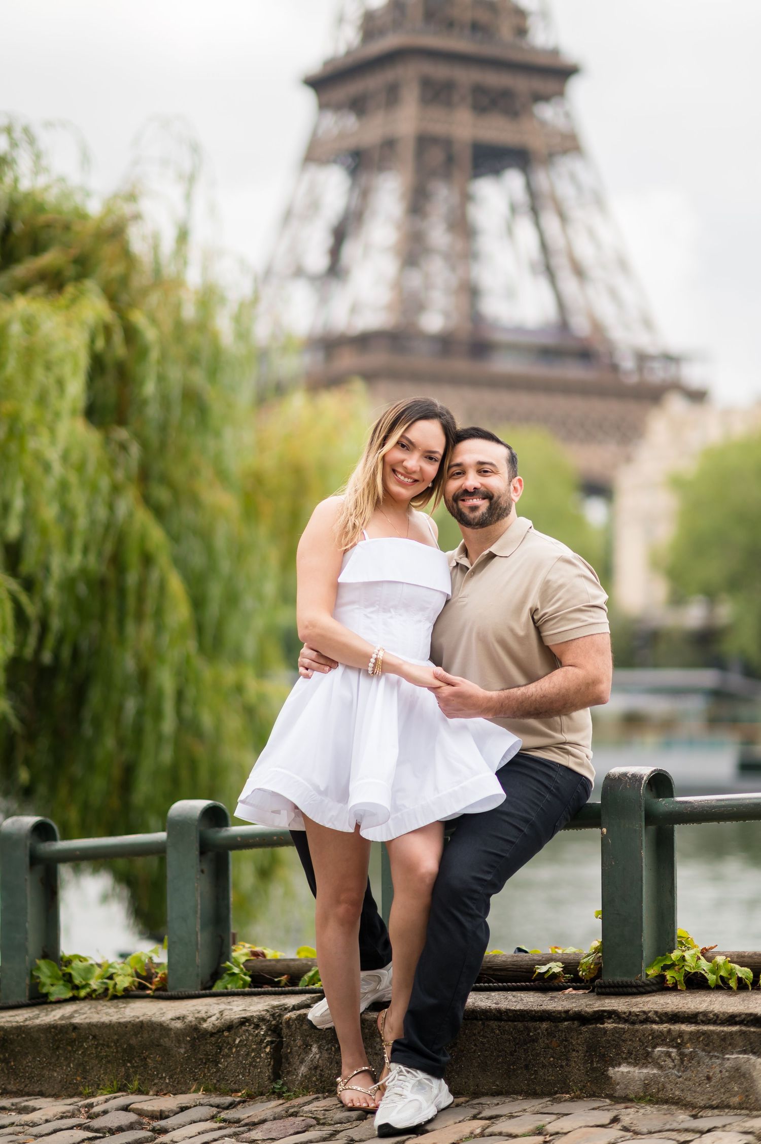 Romantic couple sits on stone wall near Eiffel Tower in Paris during springtime with green willows in background.
