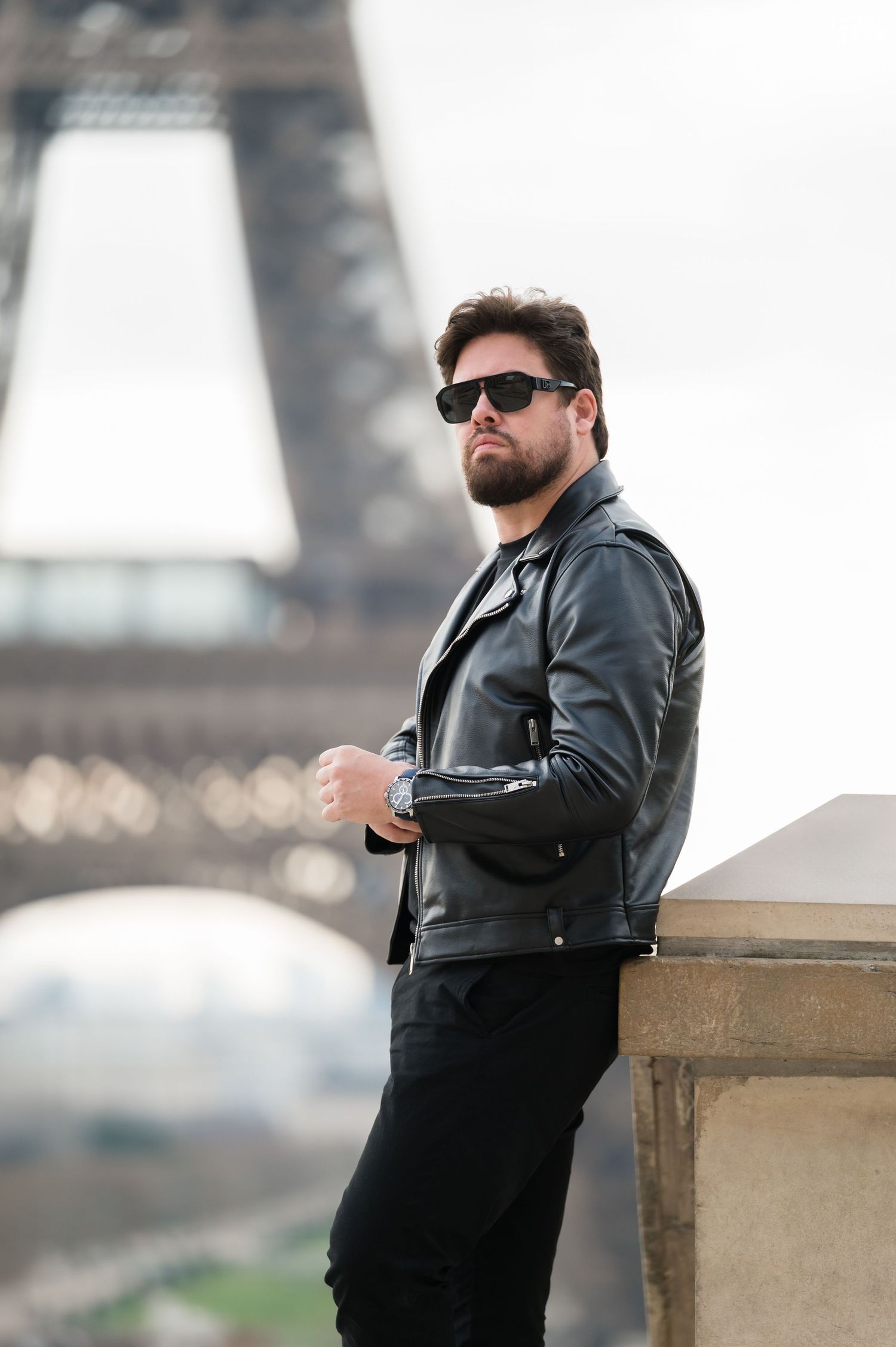 A stylish man in a black leather jacket and sunglasses poses confidently near the Eiffel Tower in Paris.
